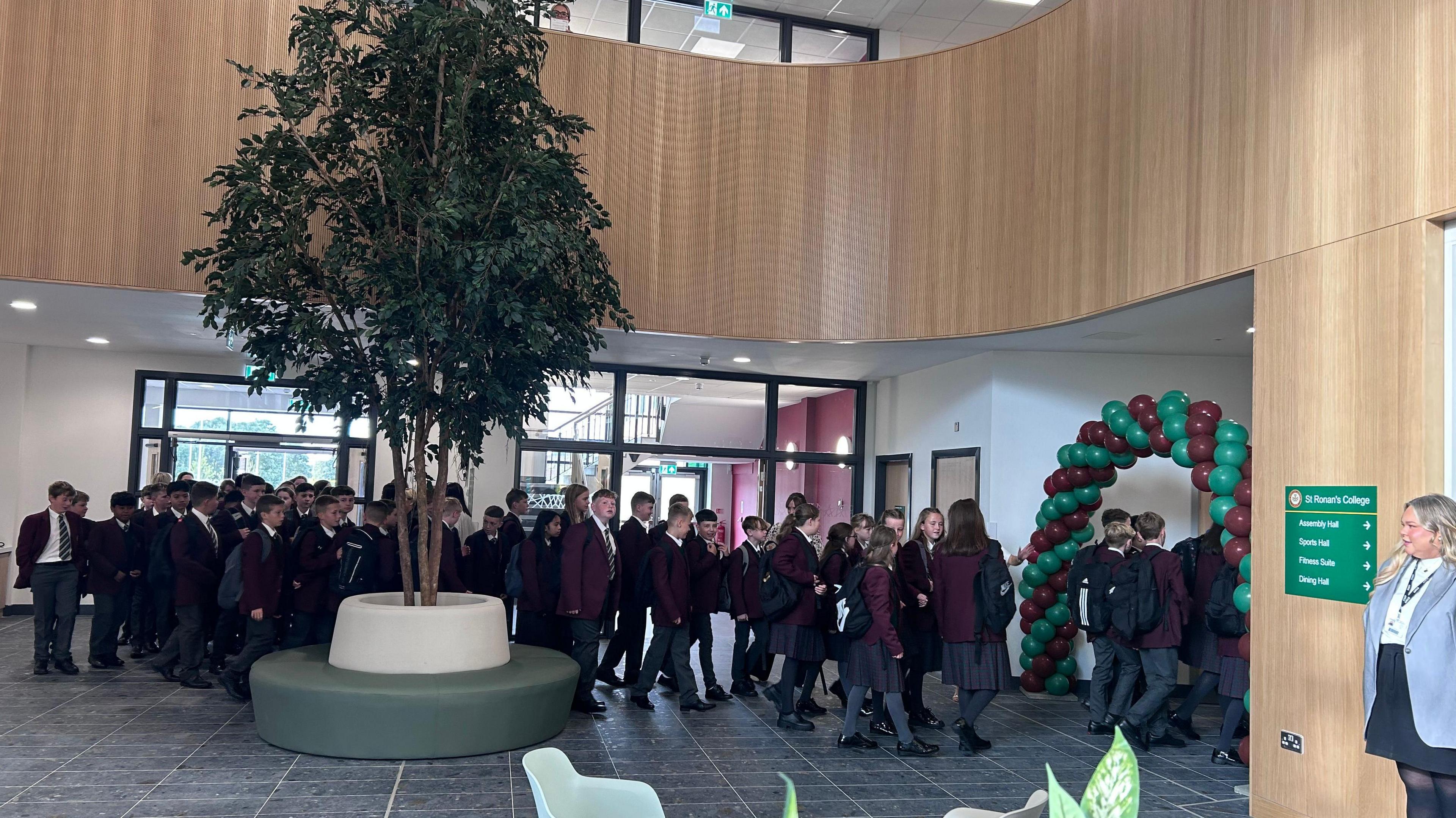 The reception area of St Ronan's. It's a large atrium with wooden panelling around the upstairs. A green and white seating area is in the middle of the downstairs with a large tree in the middle of it. Pupils are walking in a line through a green and purple balloon arch.