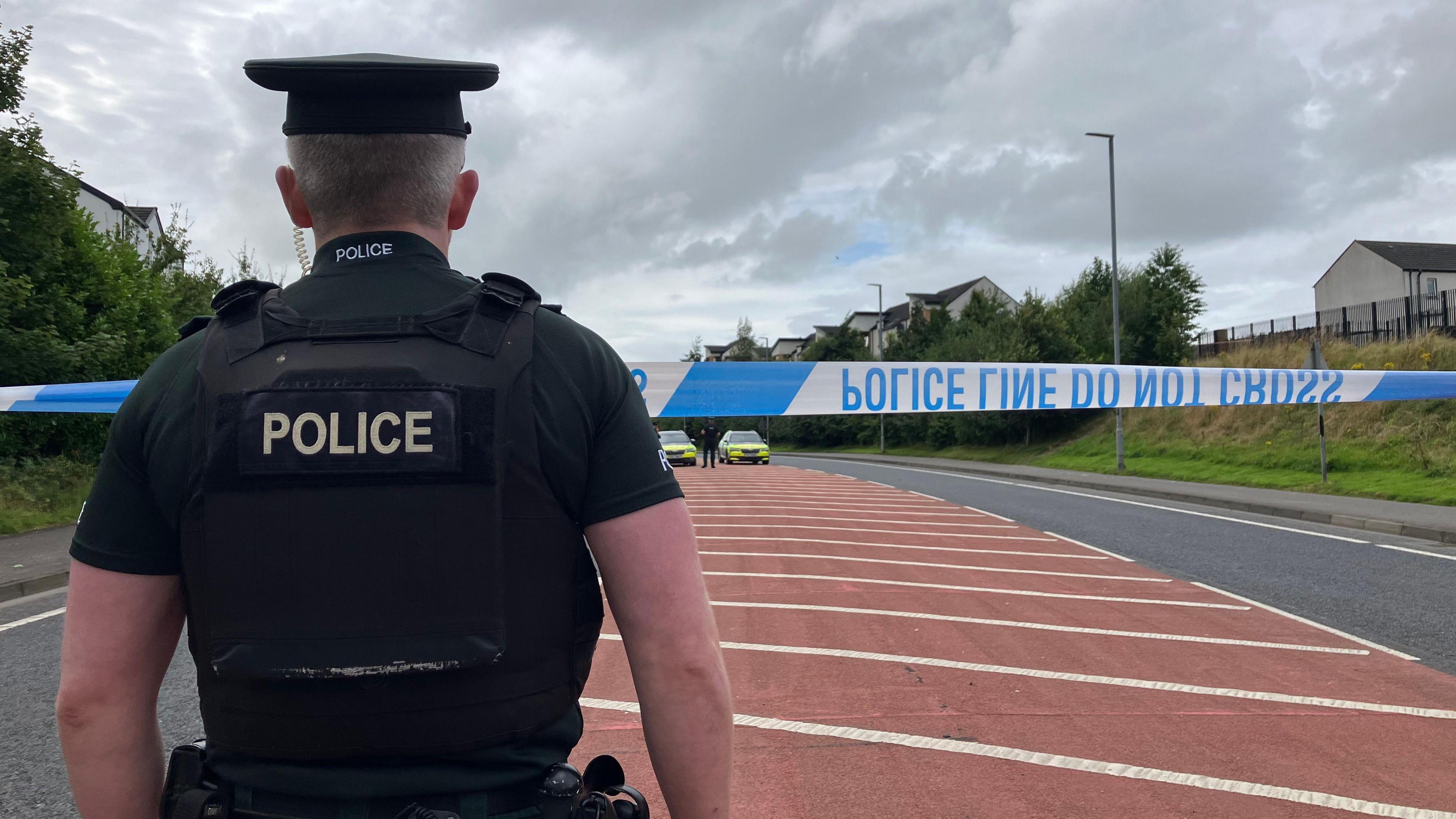 A police officer is in full uniform with his back toward the camera.
He is standing near a police cordon and a two police vehicles can be seen at the far end of a closed off road. A grassy embankment, some houses and trees line either side of the road.
