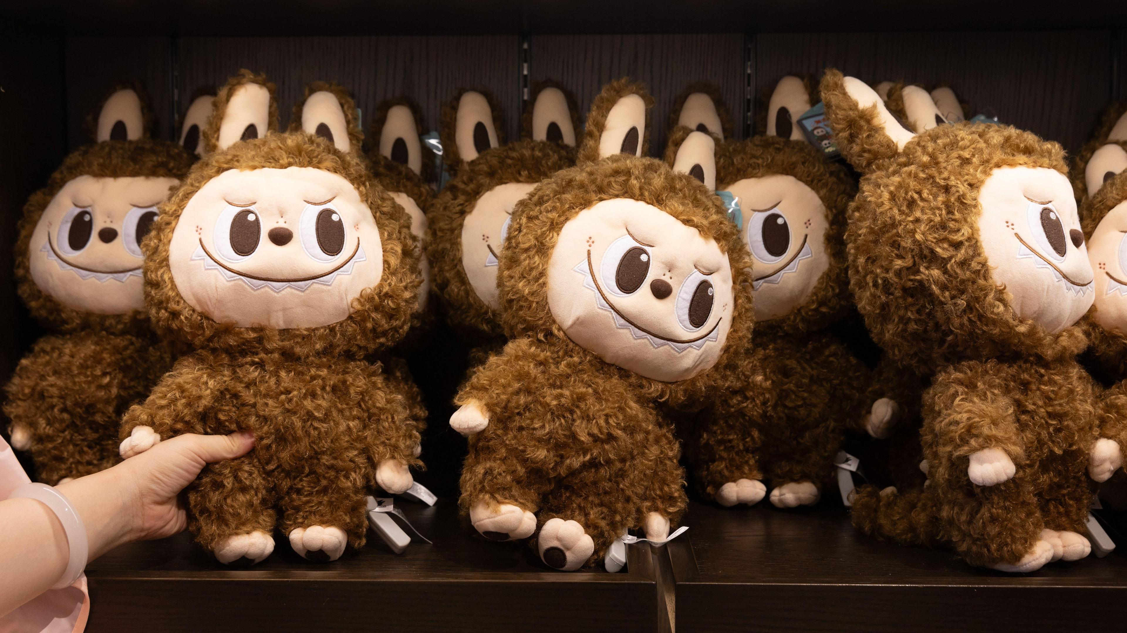 A shopper reaches for a brown furry Labubu doll, which is on a shelf with lots of other identical dolls. They all have brown fur, a light pink face and ears poking above their heads. They have big eyes and a smile.