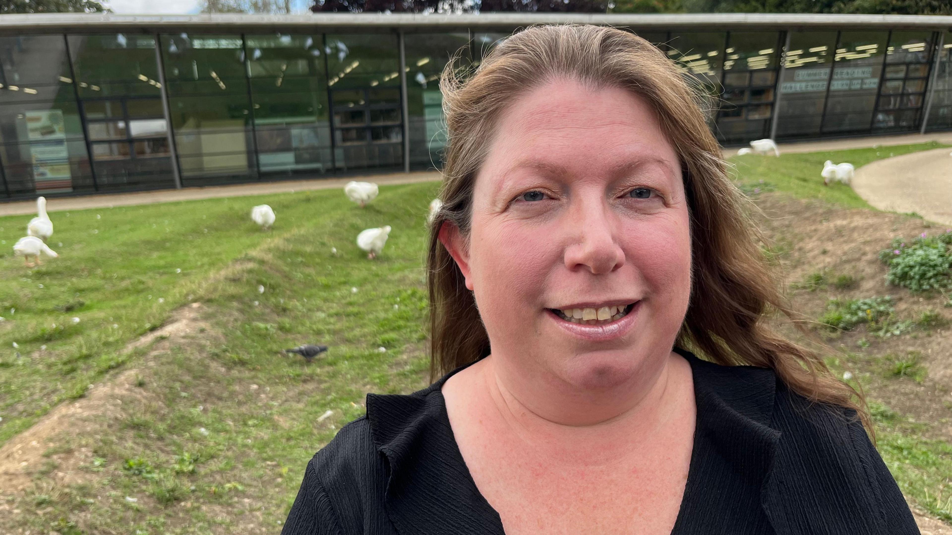 Chrissie Fadipe, 45, has shoulder length brown hair swept back over her head, she has a black V-necked top on and behind her is a public library and on ridged grass there are several white geese with orange bills and matching coloured feet.