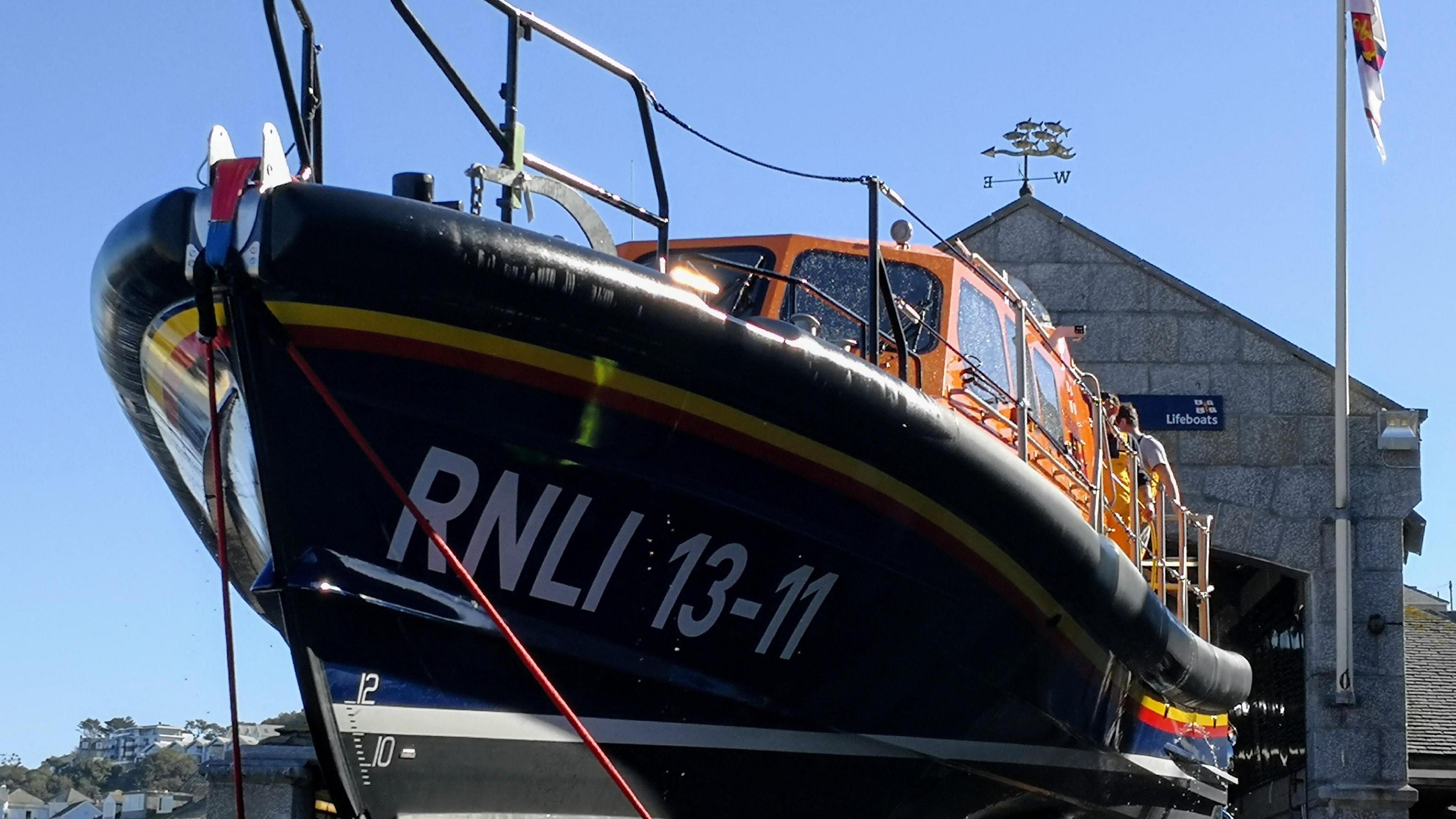 Close up shot of a RNLI vessel. It is orange and blue. Two crew members are on the boat. RNLI 13-11 is written on the side of the boat in large white letters and numbers. It is a sunny day.