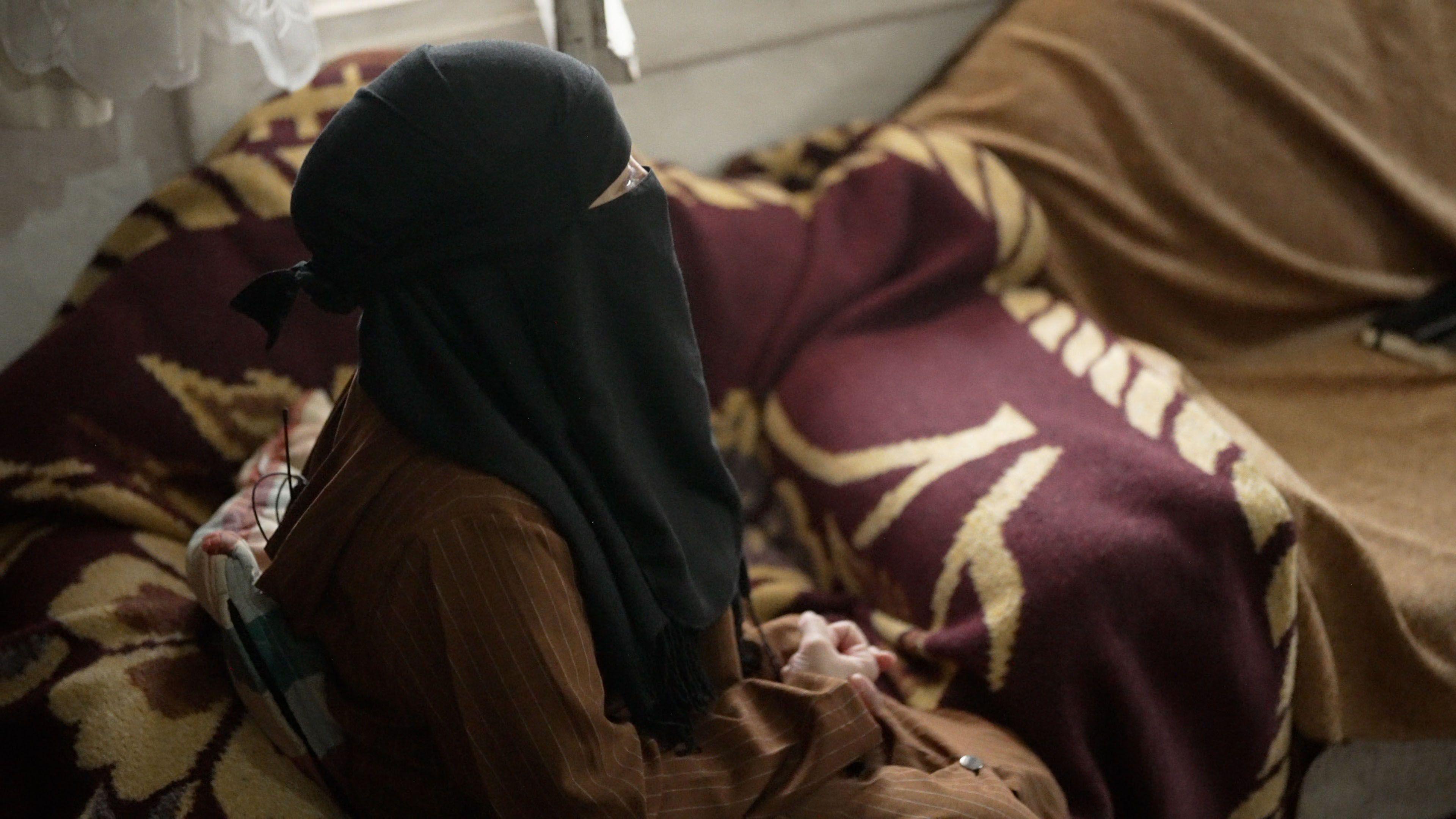 Profile of a woman sitting on a sofa. She is wearing a brown, long-sleeved dress with thin white stripes and a black headscarf which covers her face. Only part of one eye can be seen.