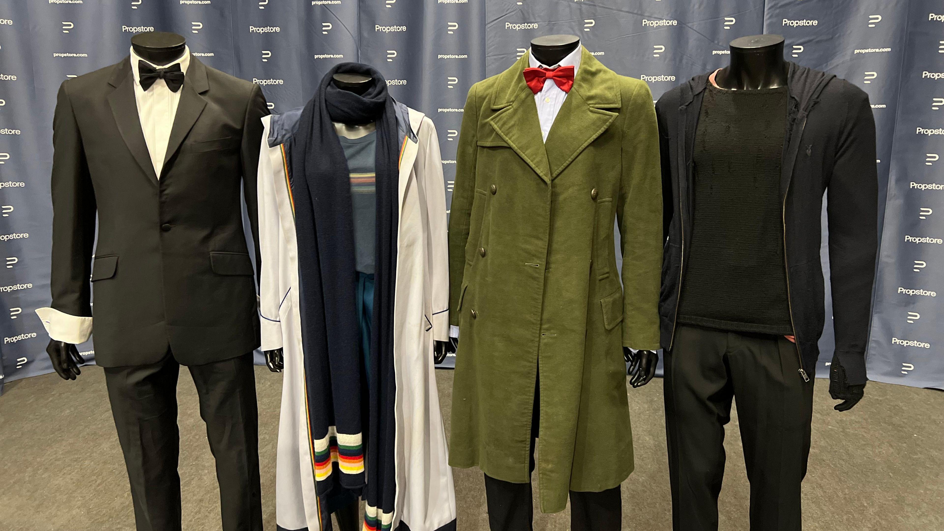 Headless mannequins wearing costumes are stood together in a line. The first has a tuxedo used by David Tennant, the second is wearing a costume used by Jodie Whittaker, a third is one used by Matt Smith and a fourth is a Peter Capaldi costume.