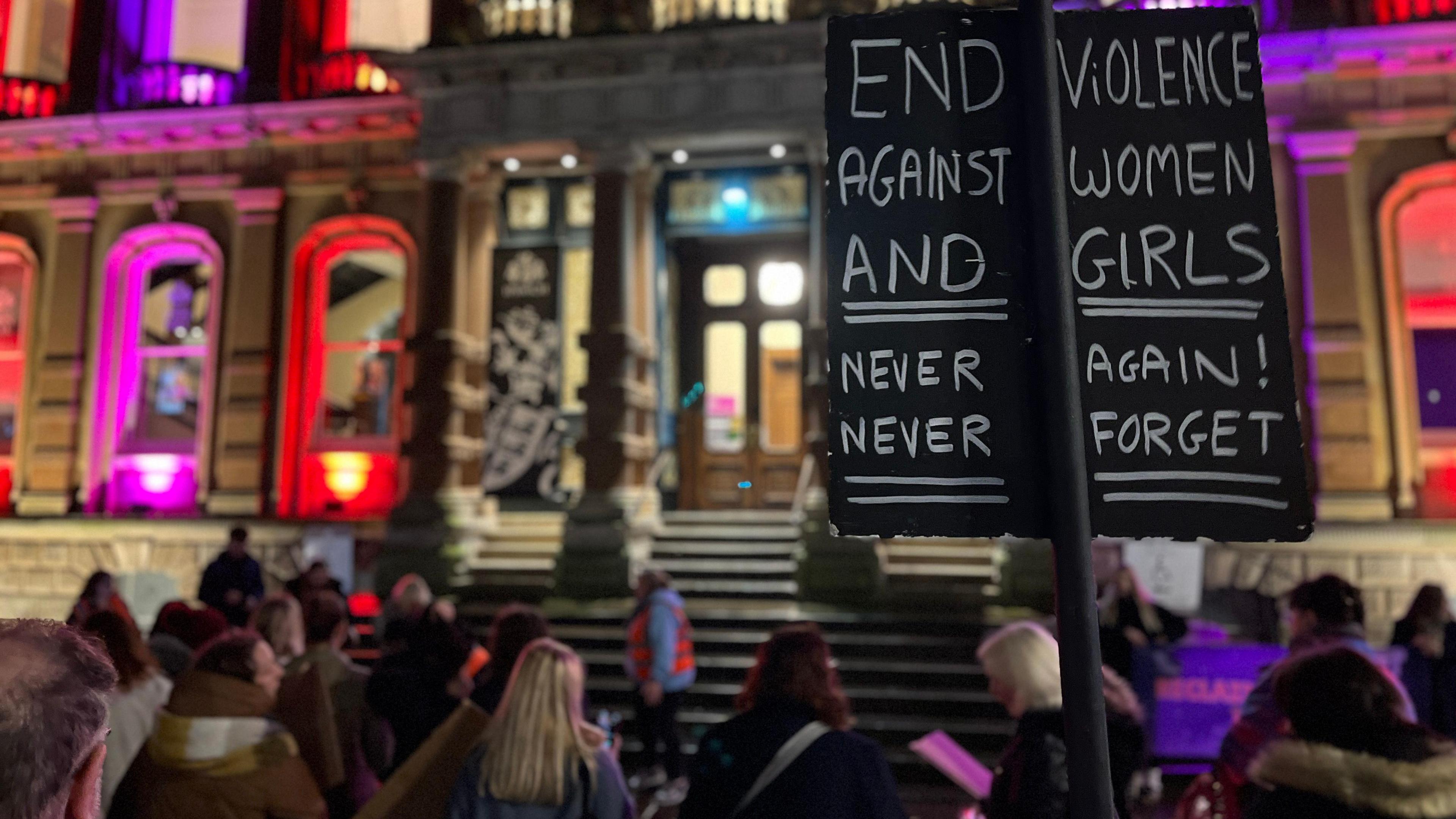 A crowd outside Ipswich town hall at night. In the foreground is a placard which reads End violence against women and girls. Never again, never forget