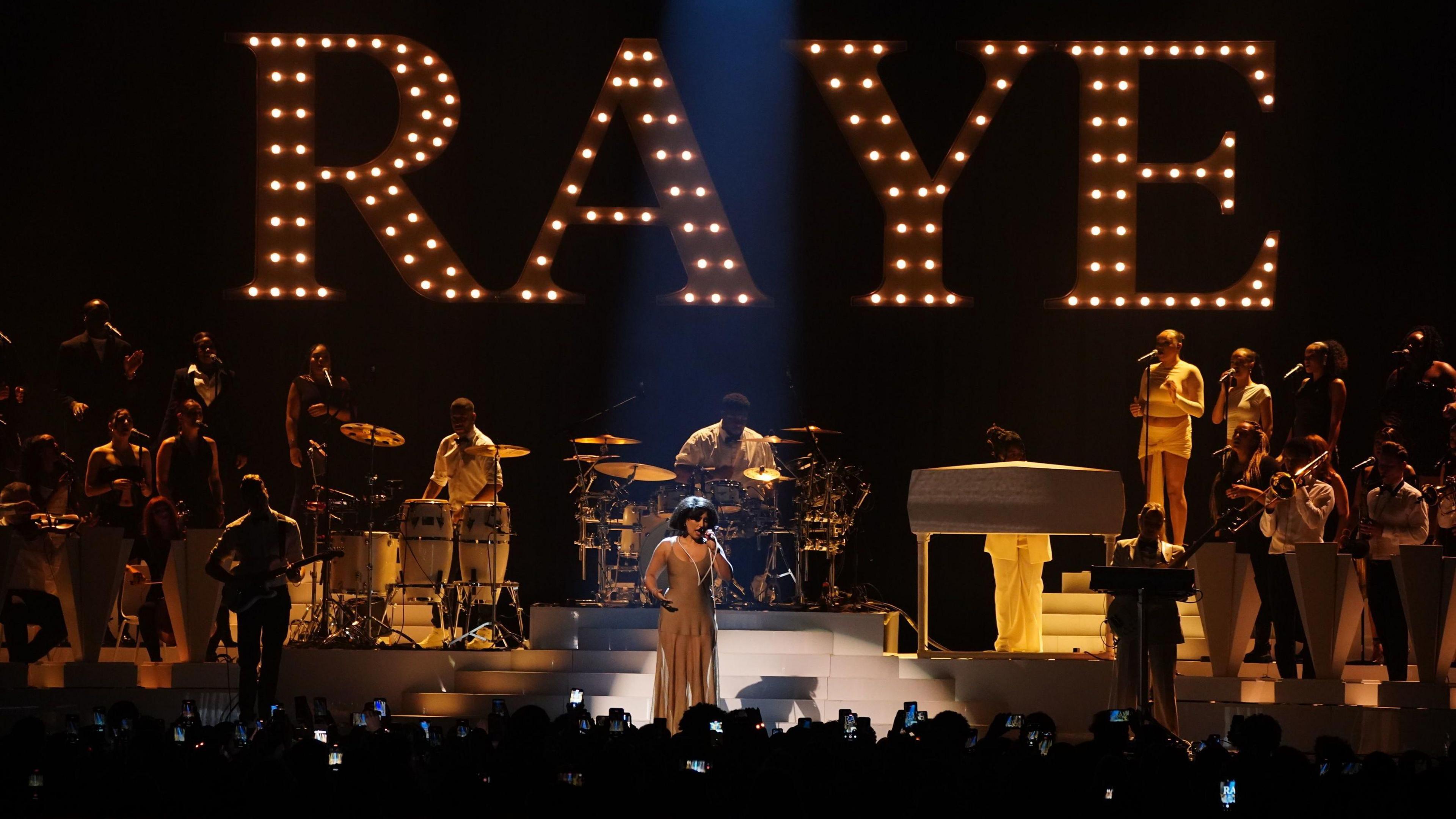 Raye performing on stage with illuminated letters spelling out Raye behind her