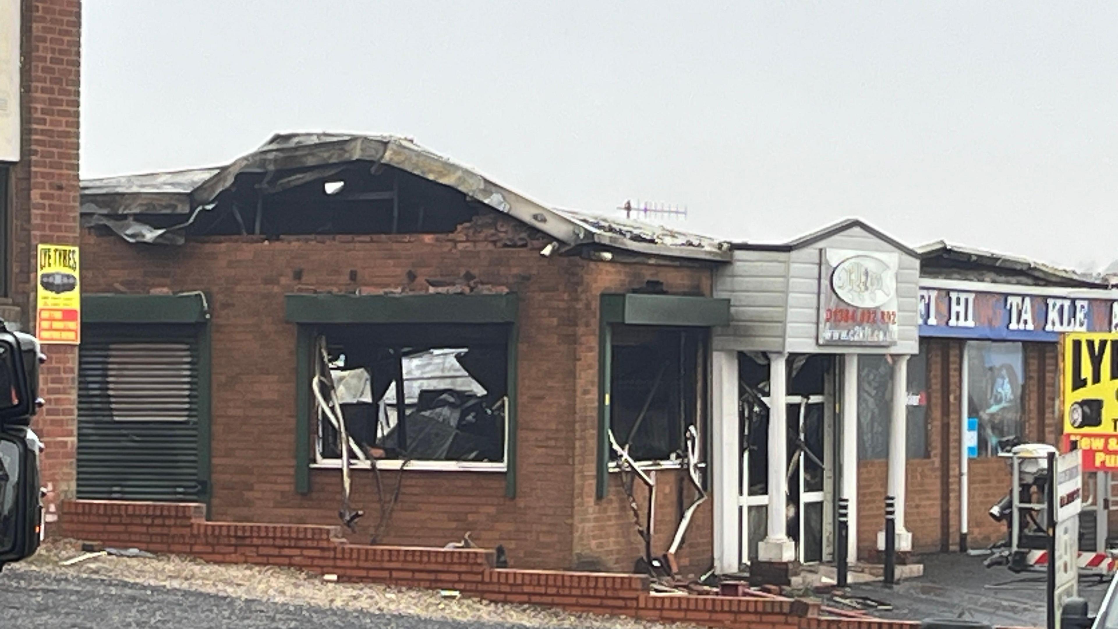 A red brick commercial building pictured from across the street in the daytime. The building is blackened in places from fire damage and there is no glass in the windows.