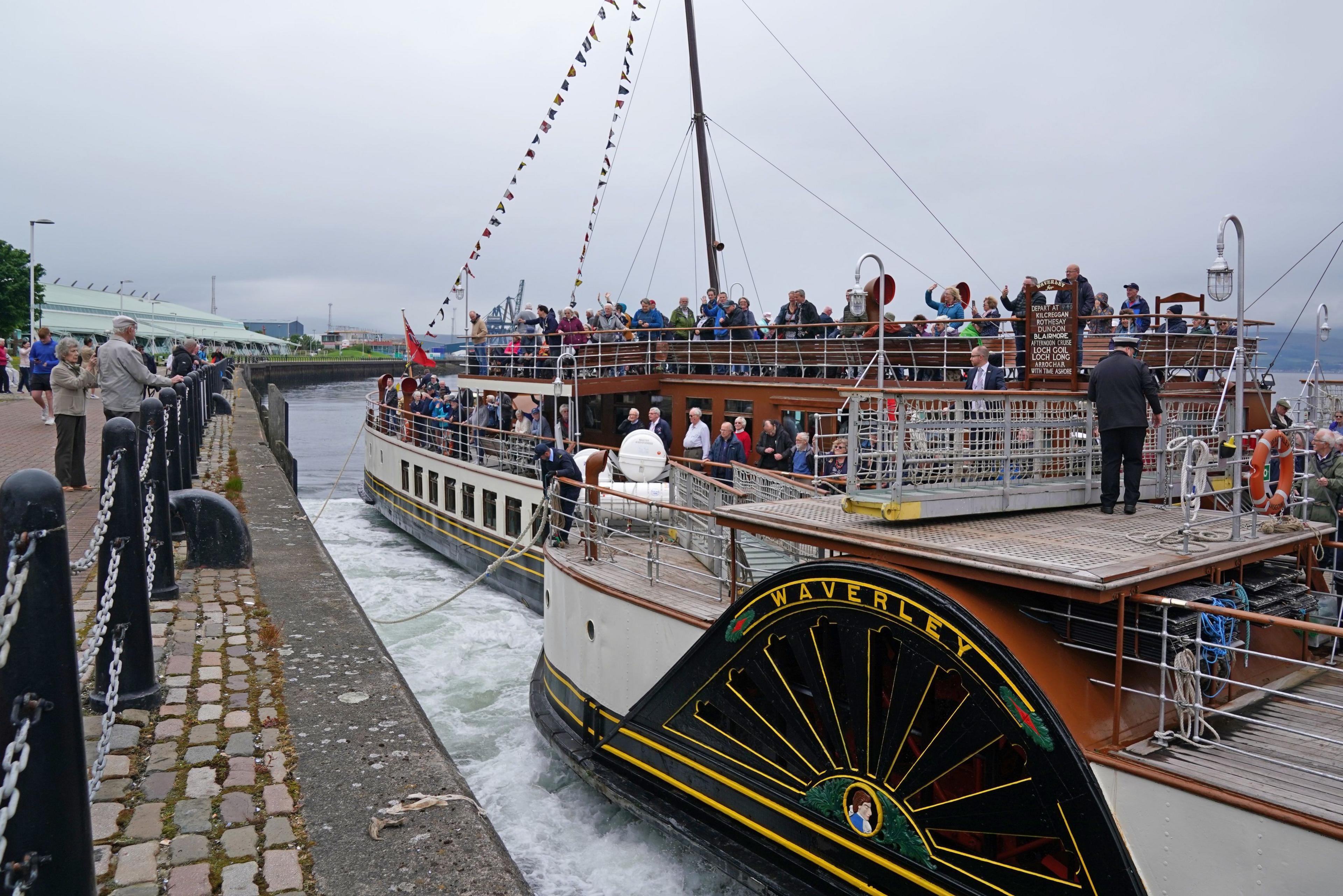 A paddle steamer filled with people, with some waving at the those on the dock.