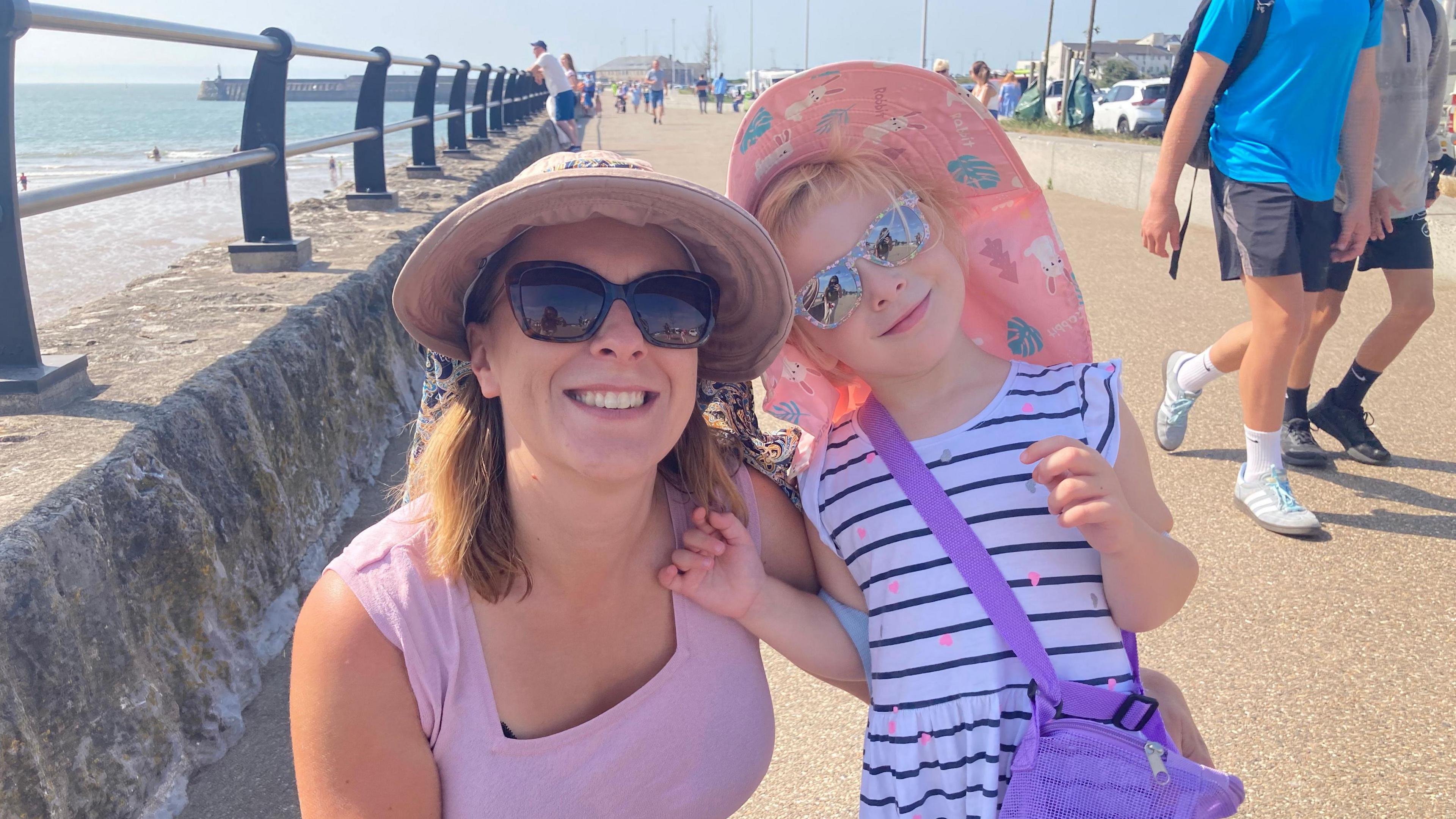 Marta ac Ella Adams mewn hetiau a sbectolau haul ar y prom ym Mhorthcawl - dynes ifanc gwallt brown mewn fest lliw lelog a merch fach bedair oed gyda gwallt melyn mewn ffrog gwyn a streipiau glas tywyll