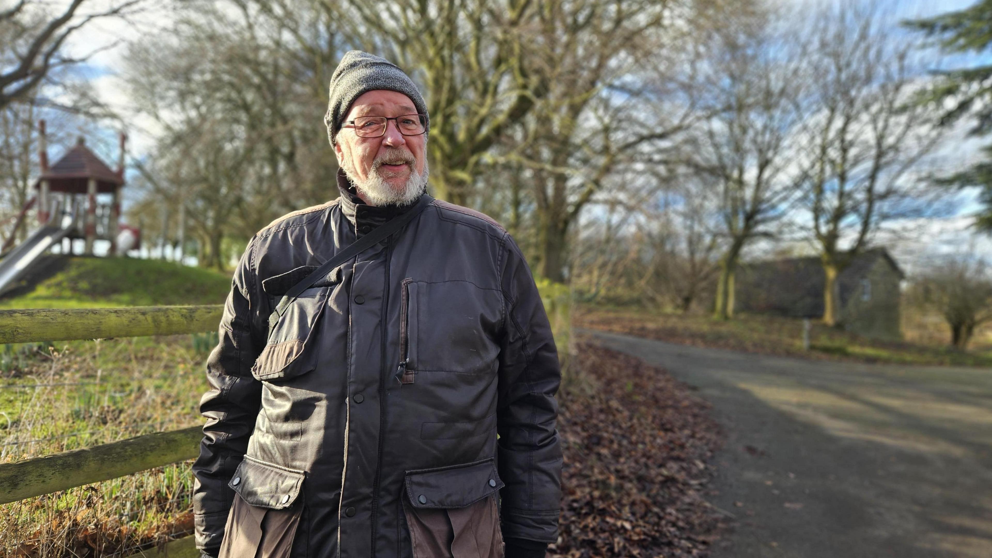 An older man with a white beard and wearing a bobble hat and coat, stands next to a wooden fence adjacent to a park. There's a public path to the other wide of him which is lined with trees, A children's play area can be seen in the background.