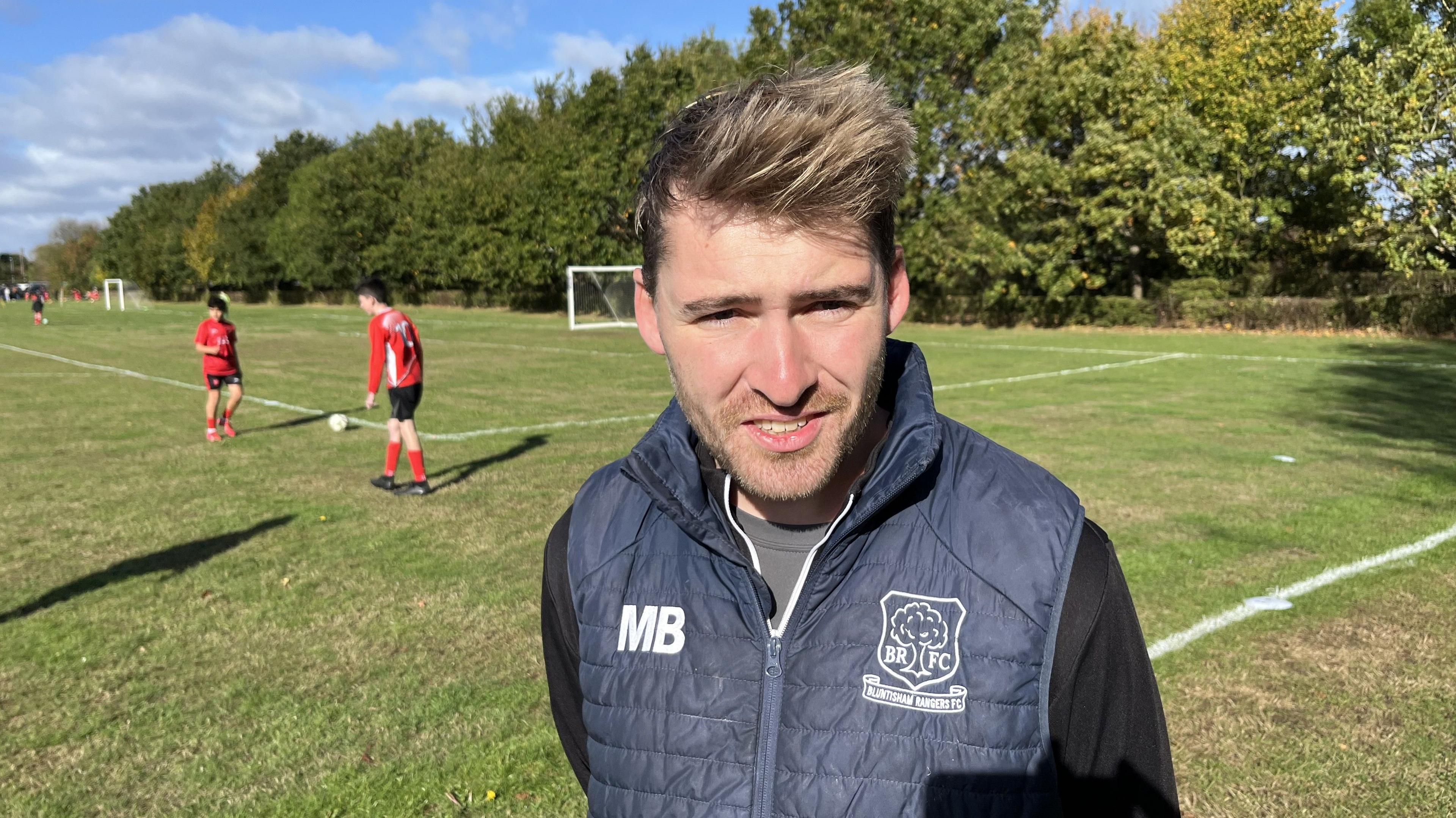 Max Brindley looks into the camera - he is on the football pitch and wearing a padded navy gilet with his initials MB on it