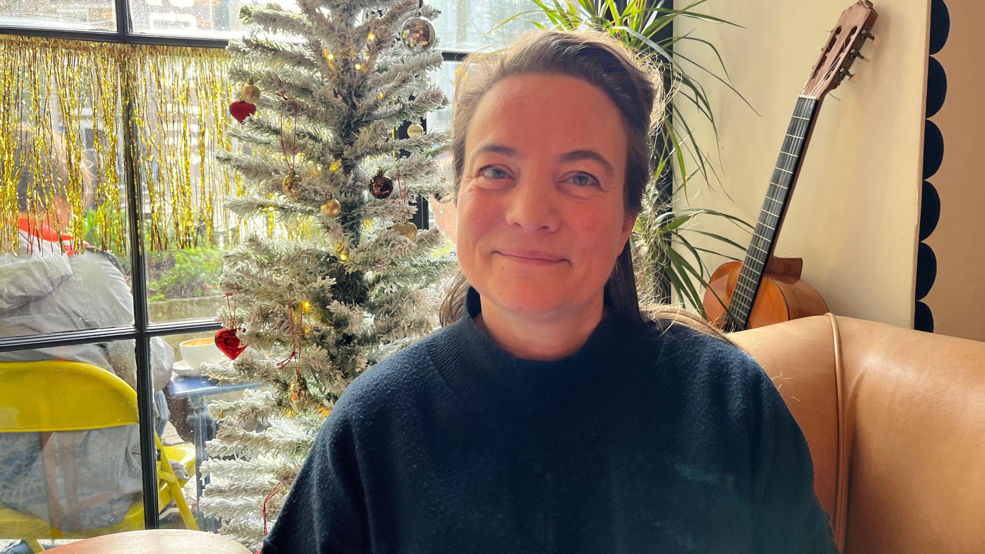 Cafe owner Lou Rose is sitting on a beige sofa with a Christmas tree behind her. Lou is wearing a dark jumper. She has brown hair and is smiling at the camera. 