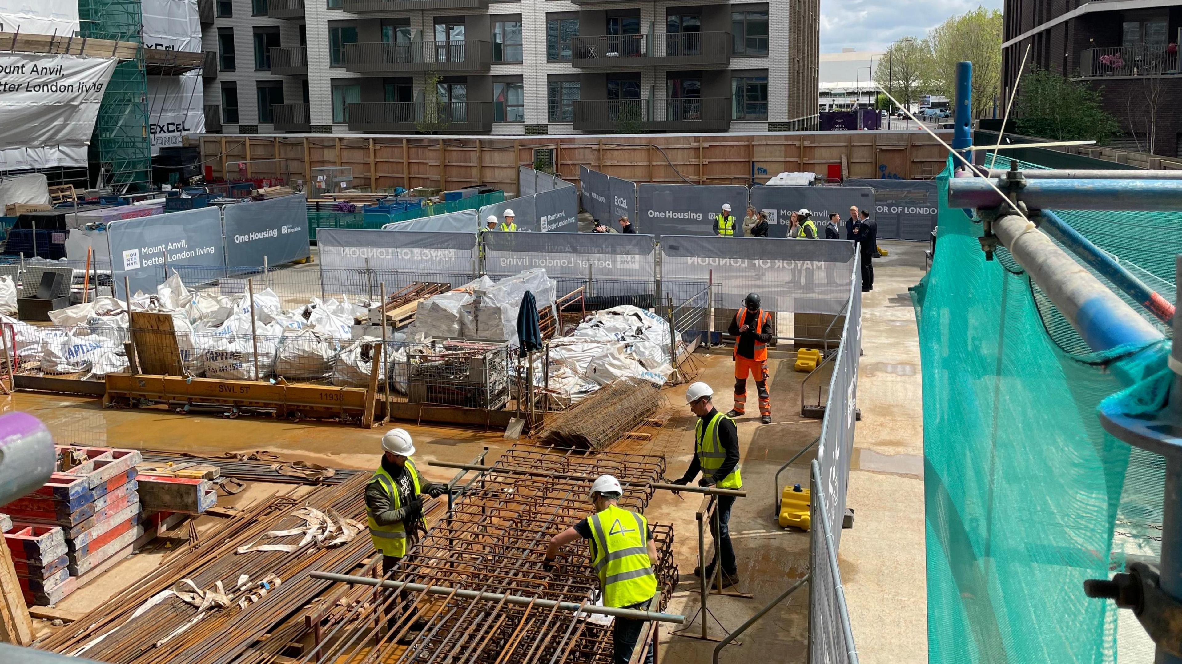 Groups of builders are handling metal grids on a building site. They are wearing high viz clothing and hard hats. In the background are more builders in high viz clothing and hard hats. A partially constructed building is also in the backgound. 