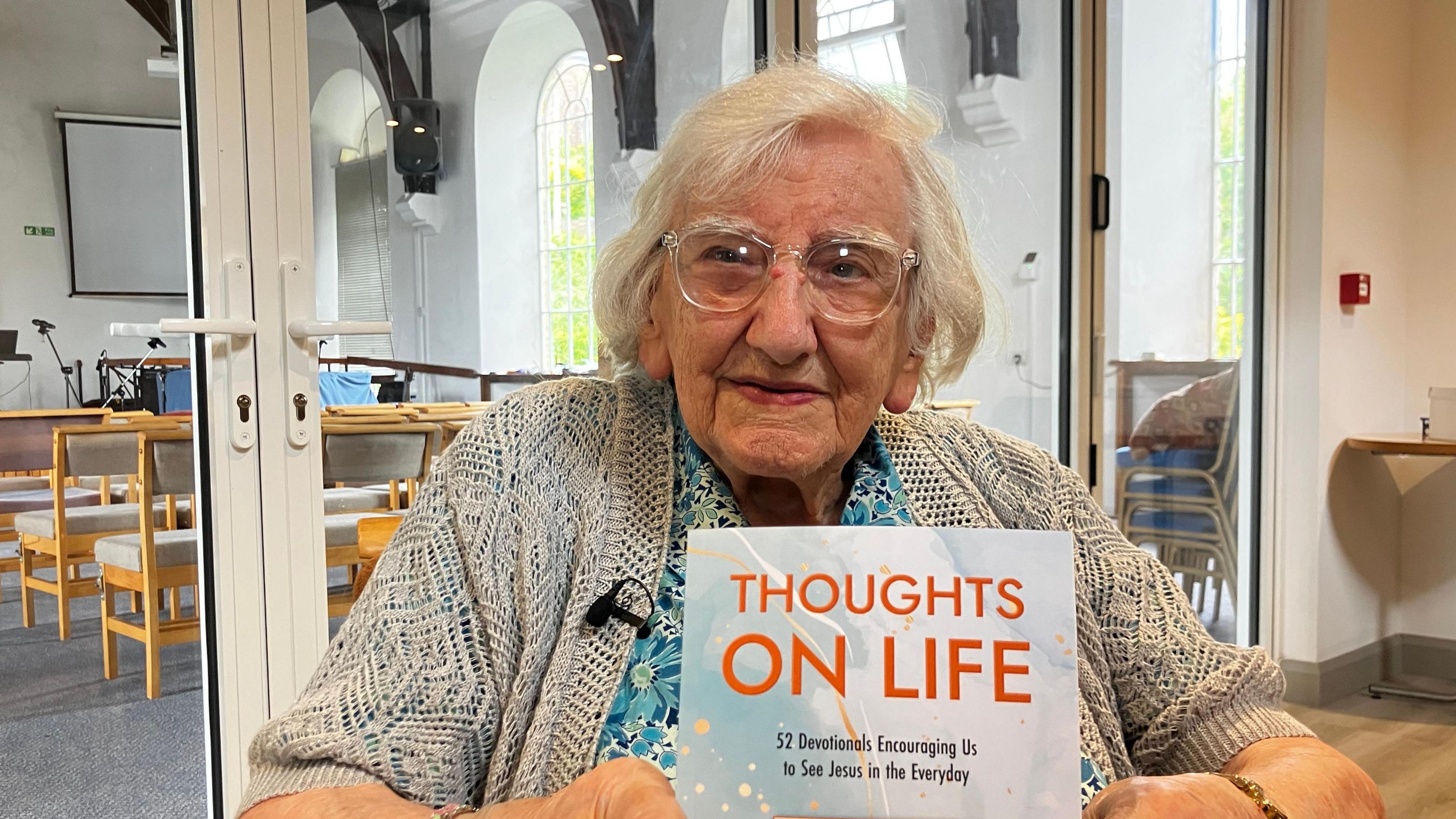 Marjorie Mitchell has short silver hair and glasses. She is wearing a blue flower-print dress and grey cardigan, sitting in church holding her book up to the camera. The title of the book is Thoughts on Life.