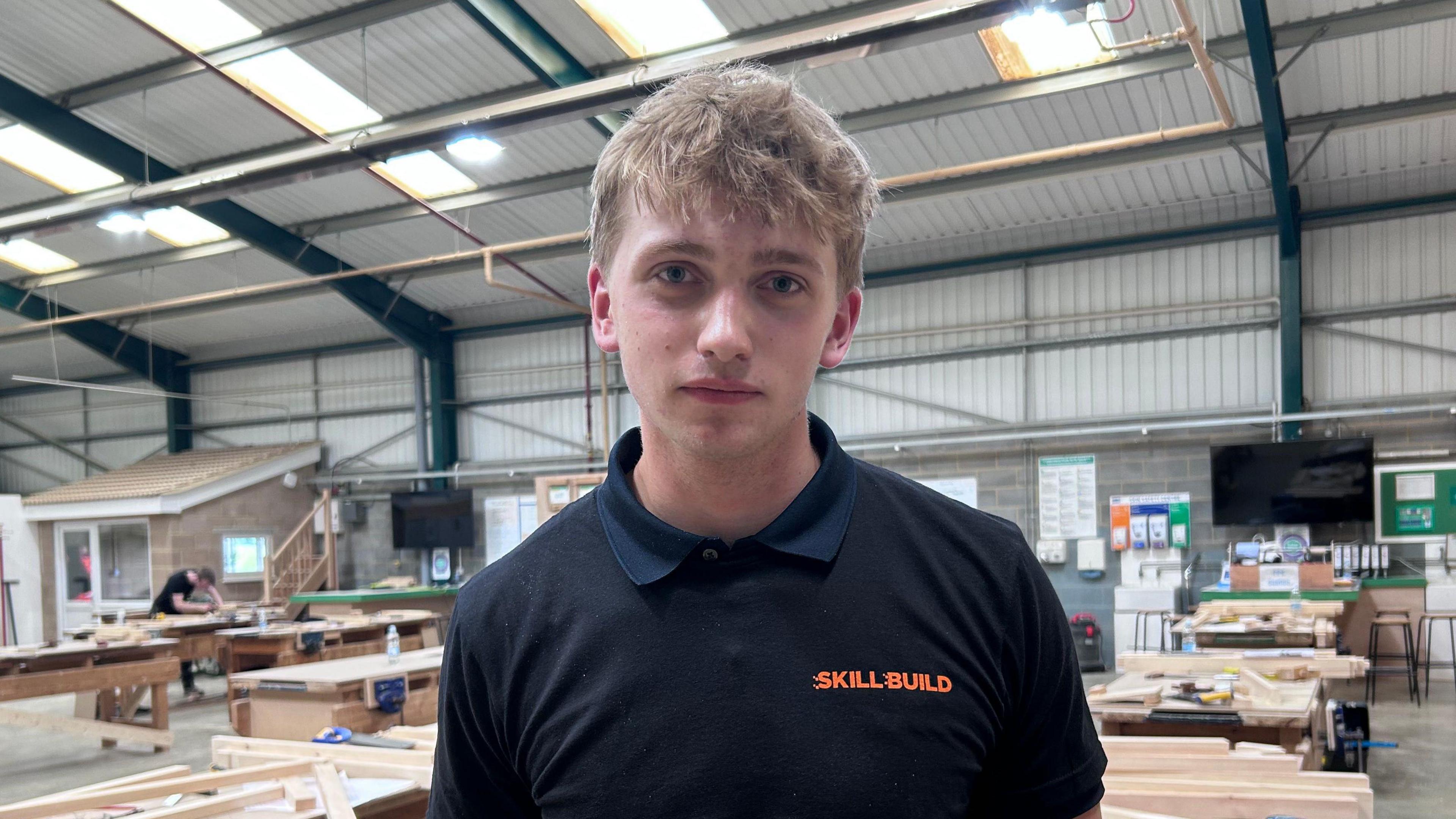 Sean with short blond hair looking at the camera and wearing a Skillbuild branded polo shirt. He is inside a large metal-framed workshop with wooden benches in the background. A person is working on a project in the distance to the left.