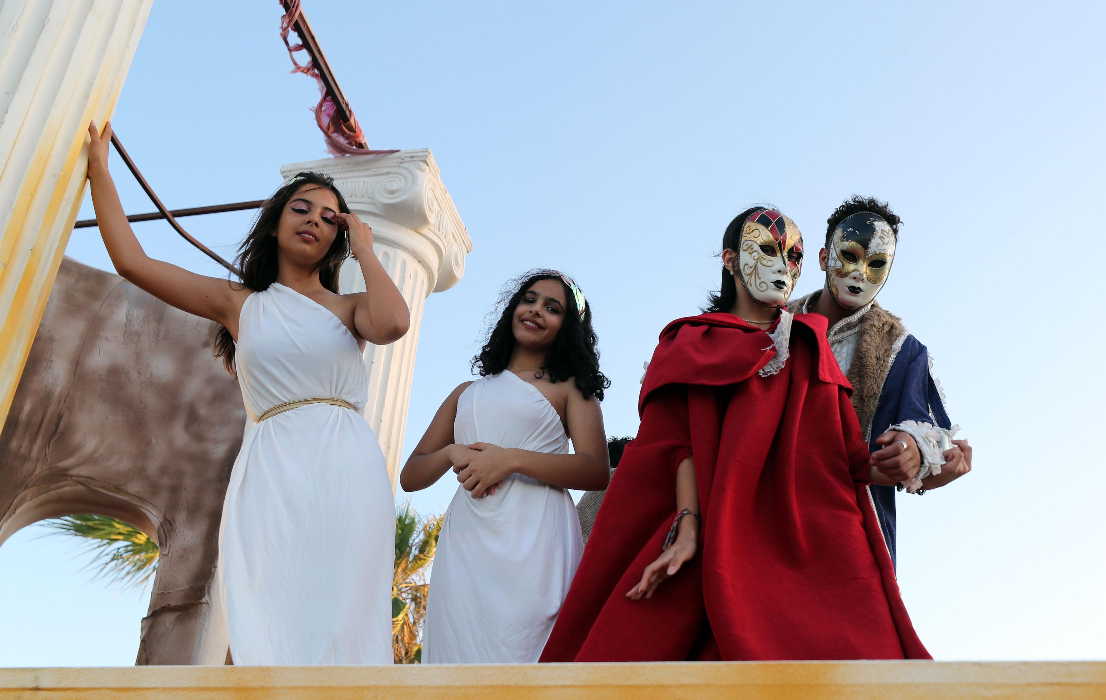 People dressed in white togas and Venetian ball attire stand on a moving float that has Greco-Roman columns in the background.
