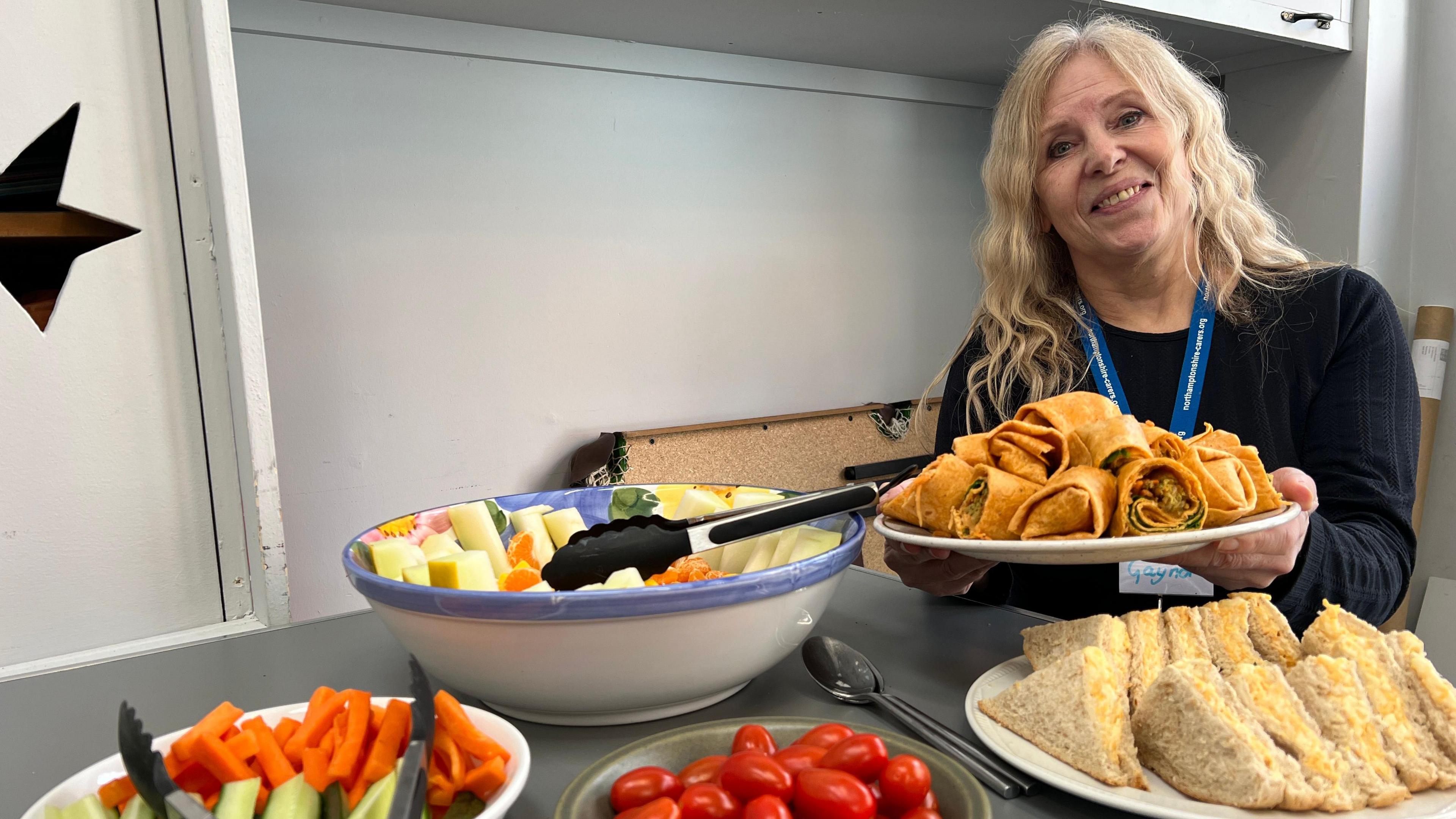 Gaynor Roberts, who works for Northamptonshire Carers, is in charge of organising the diabetes group meetings and chooses what's on the menu for lunch. She has blonde wavy hair and is wearing a black top. Gaynor is smiling and is sat a table with a plate of wraps  filled with salad and falafel in her hands. The table is also laid with a large bowl of fruit salad, orange tomatoes, as well as slices of green cumber and orange carrots. 