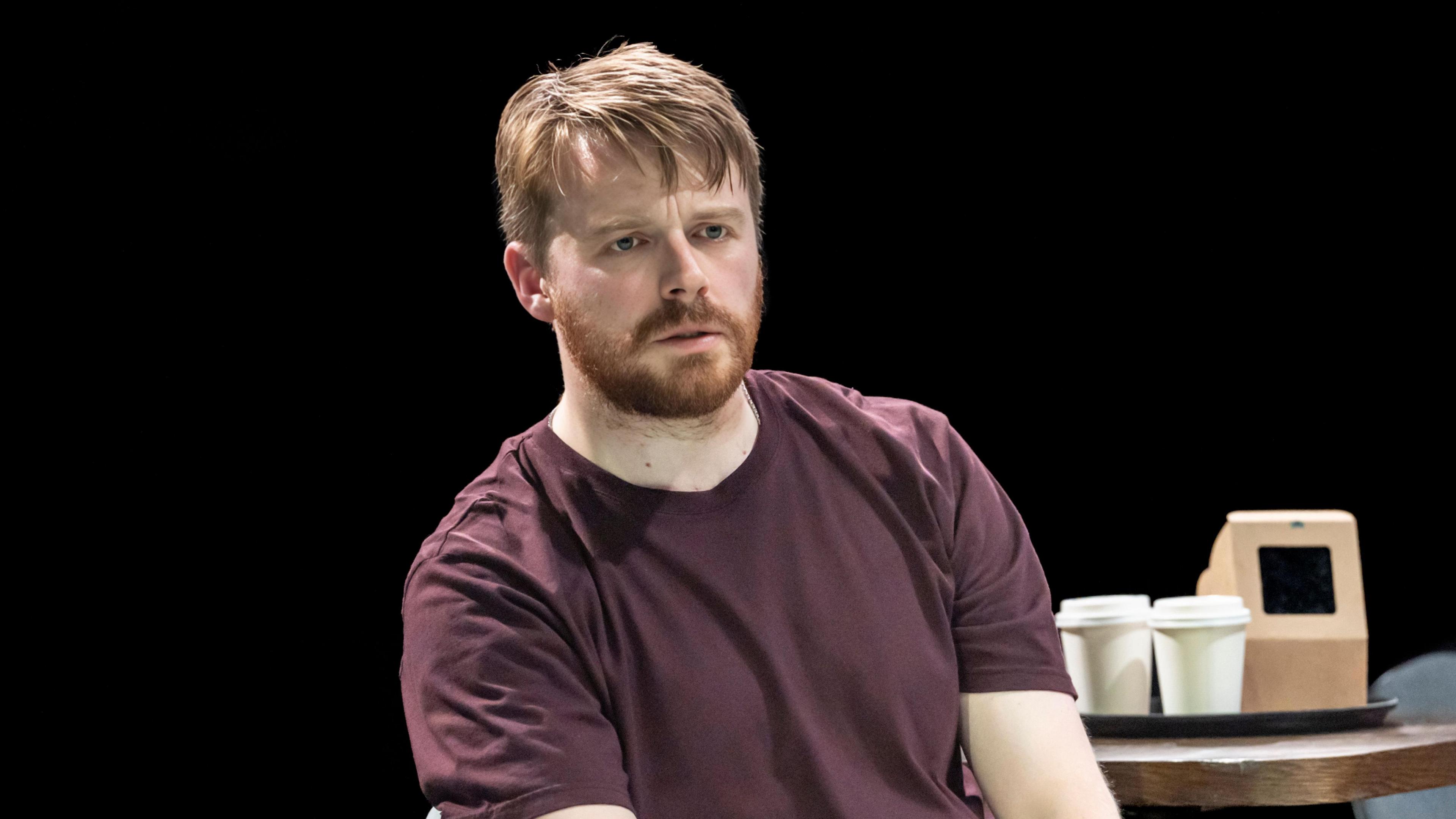 Jack Lowden sits in a chair on stage in The Fifth Step, looking concerned, and wearing a burgundy T-shirt.