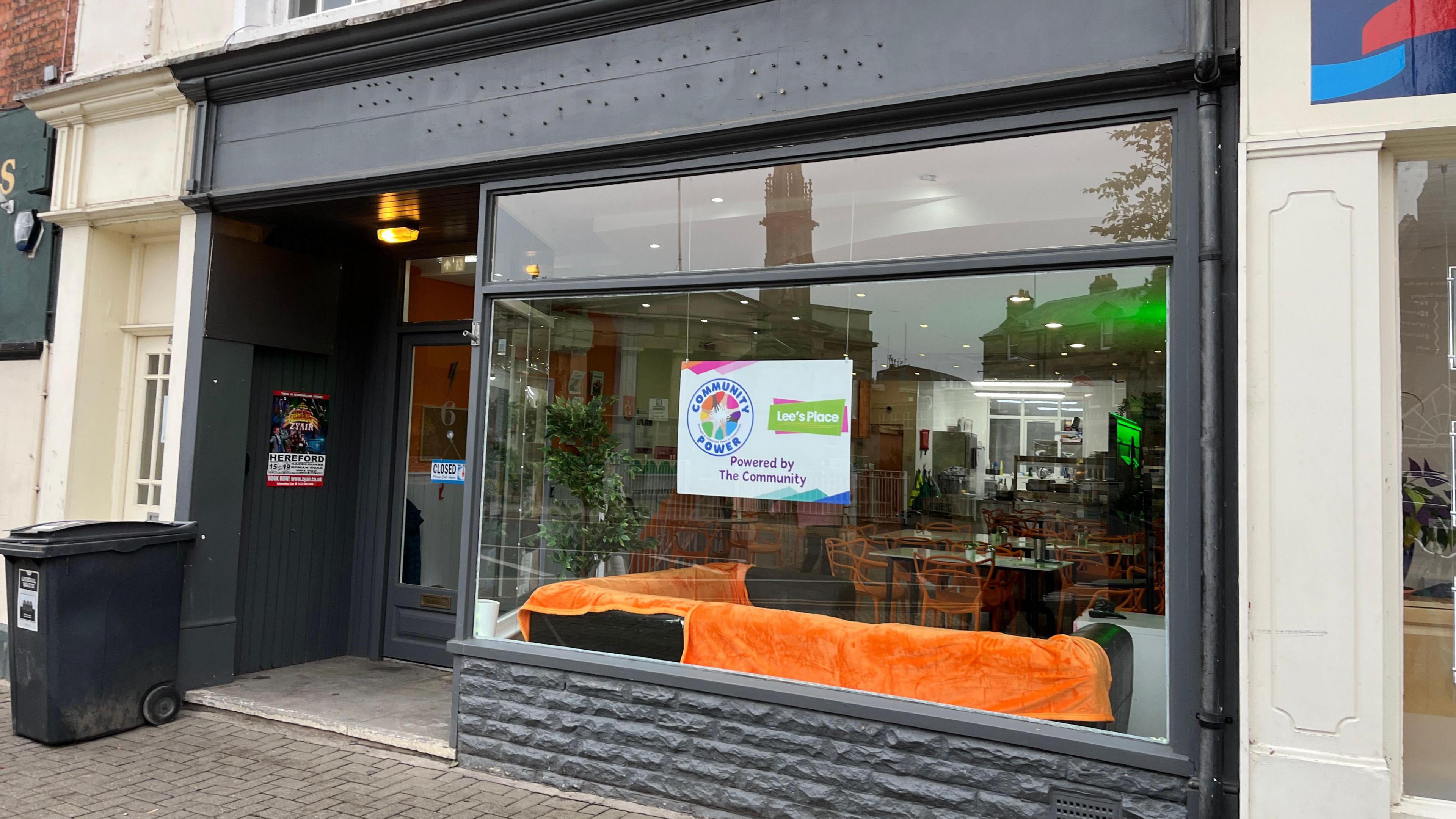 The shop frontage to the charity is painted black with a large glass window. Inside there are sofas, chairs and tables.