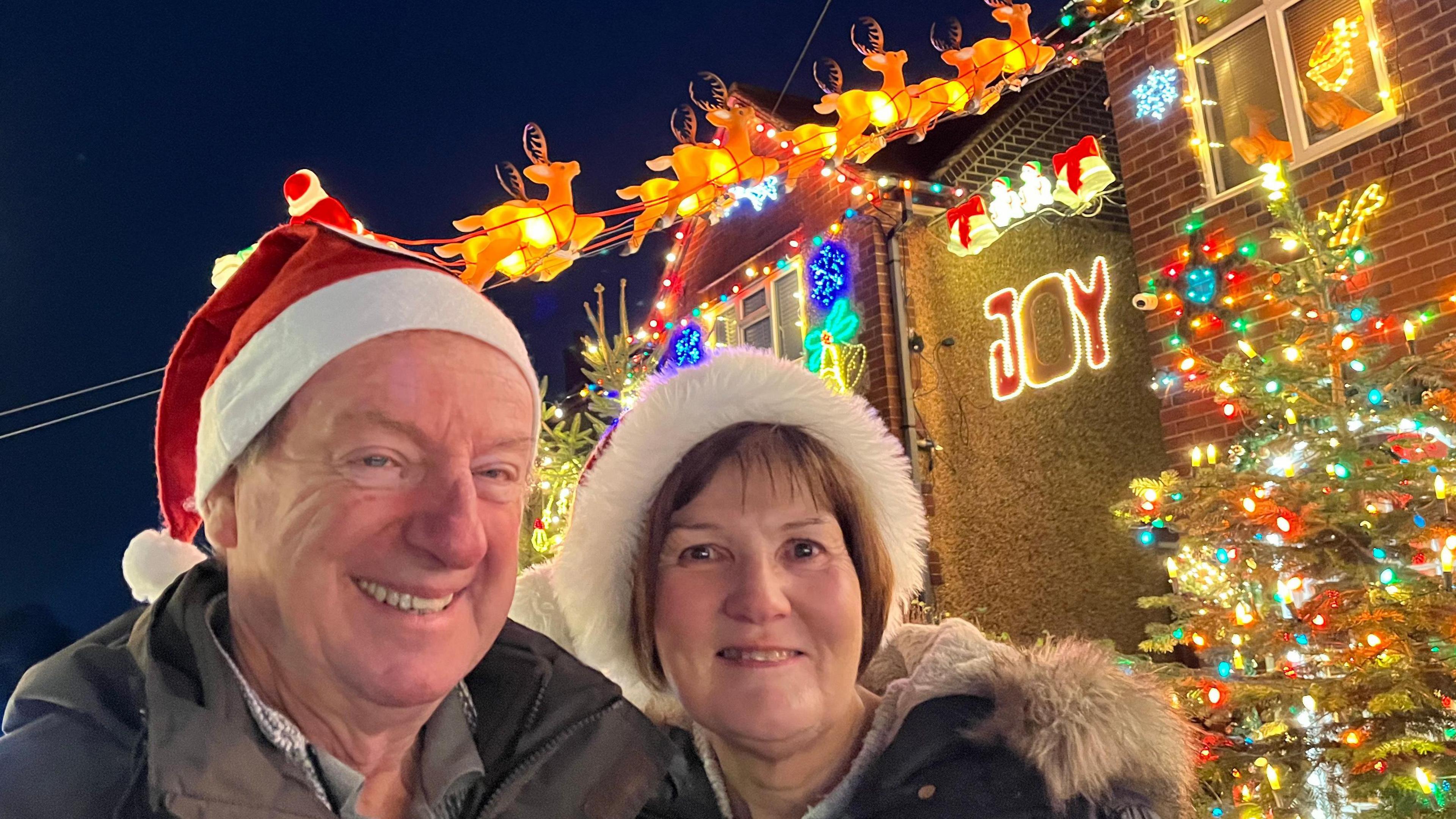 Chris and Christine stand outside a house wearing Santa hats. Behind them is a lit up sign saying "Joy", as well as a Christmas tree and reindeer decorations