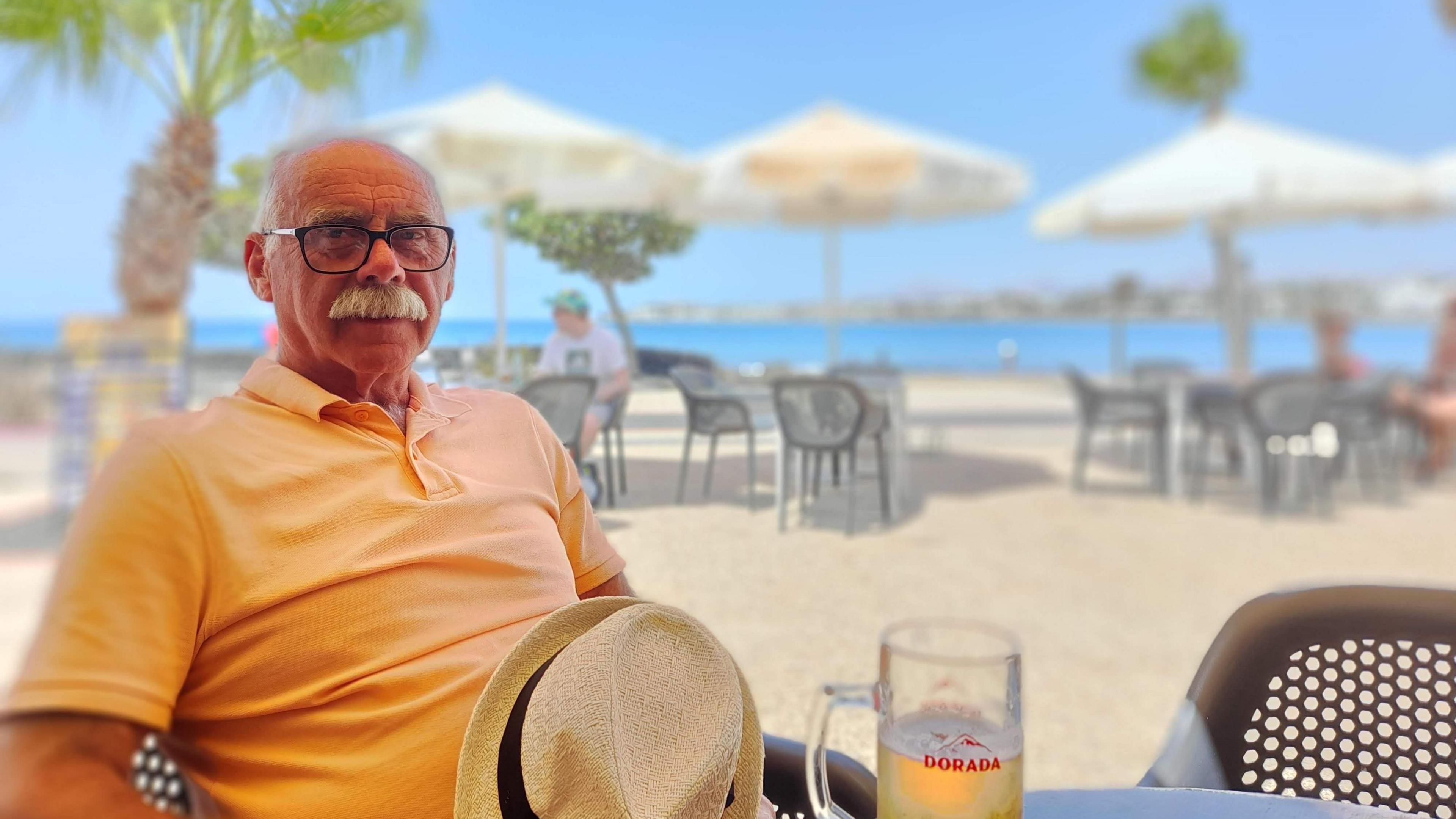 David is sat at a beach cafe with palm trees and shaded tables and chairs on the sand in the background. He is sat at a table with a glass of lager and his sun hat. He is wearing a pale orange polo shirt. He has dark-rimmed glasses and a white moustache.