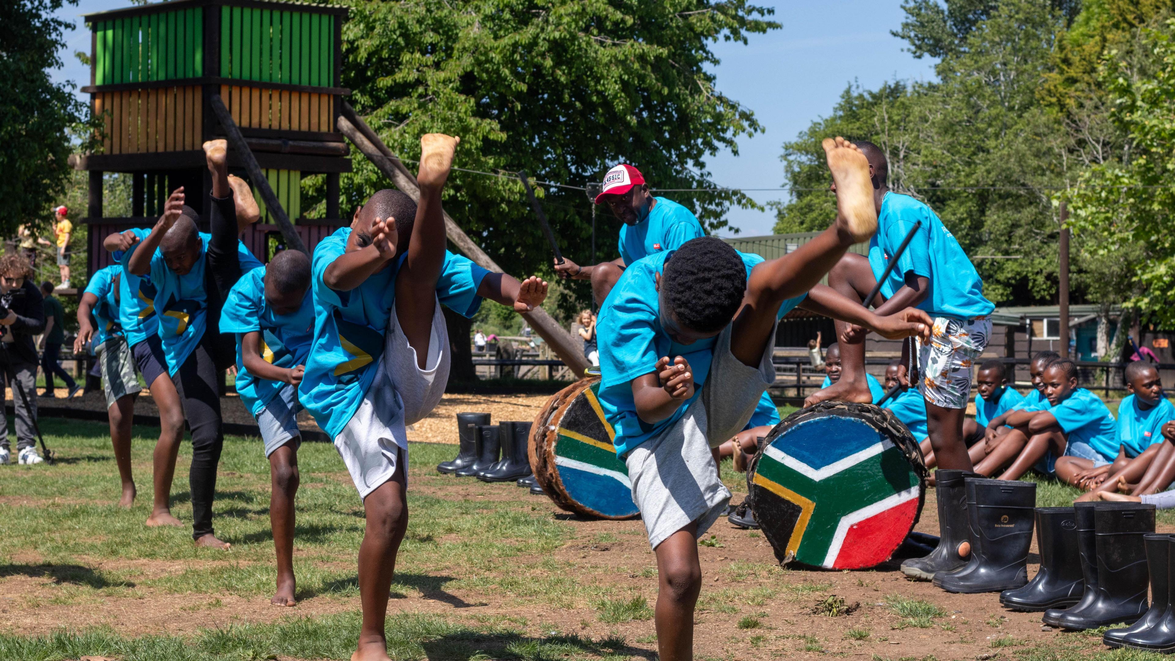 A line of boys performing. All have their legs in the air and are performing in an outdoor grassy environment.