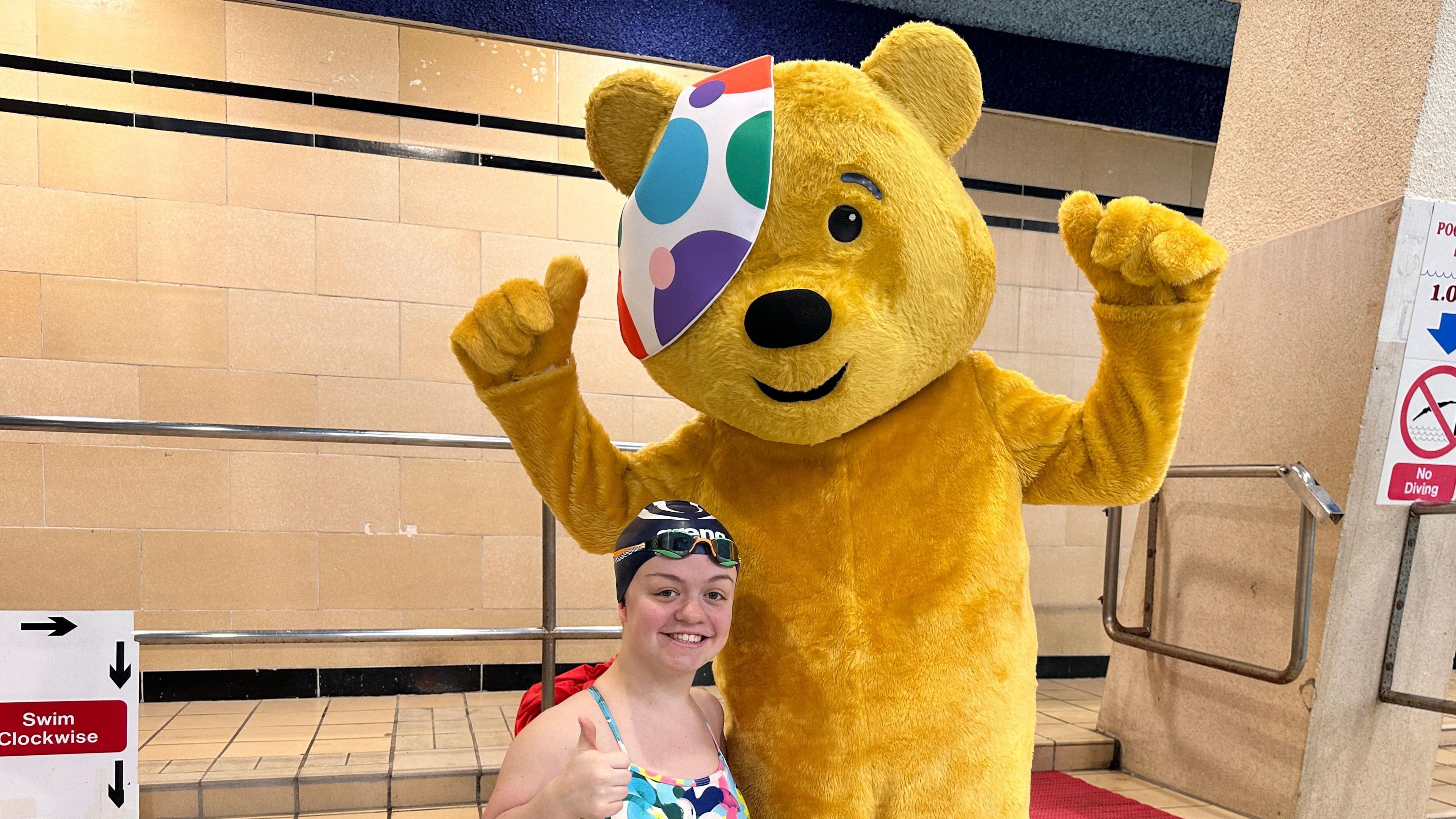 A yellow furry bear wearing a white polka dot eye patch puts his thumbs up standing next to a smiling Maisie Summer-Newton poolside. Maisie is wearing a black swimming cap, black racing goggles and a multi coloured swim suit.