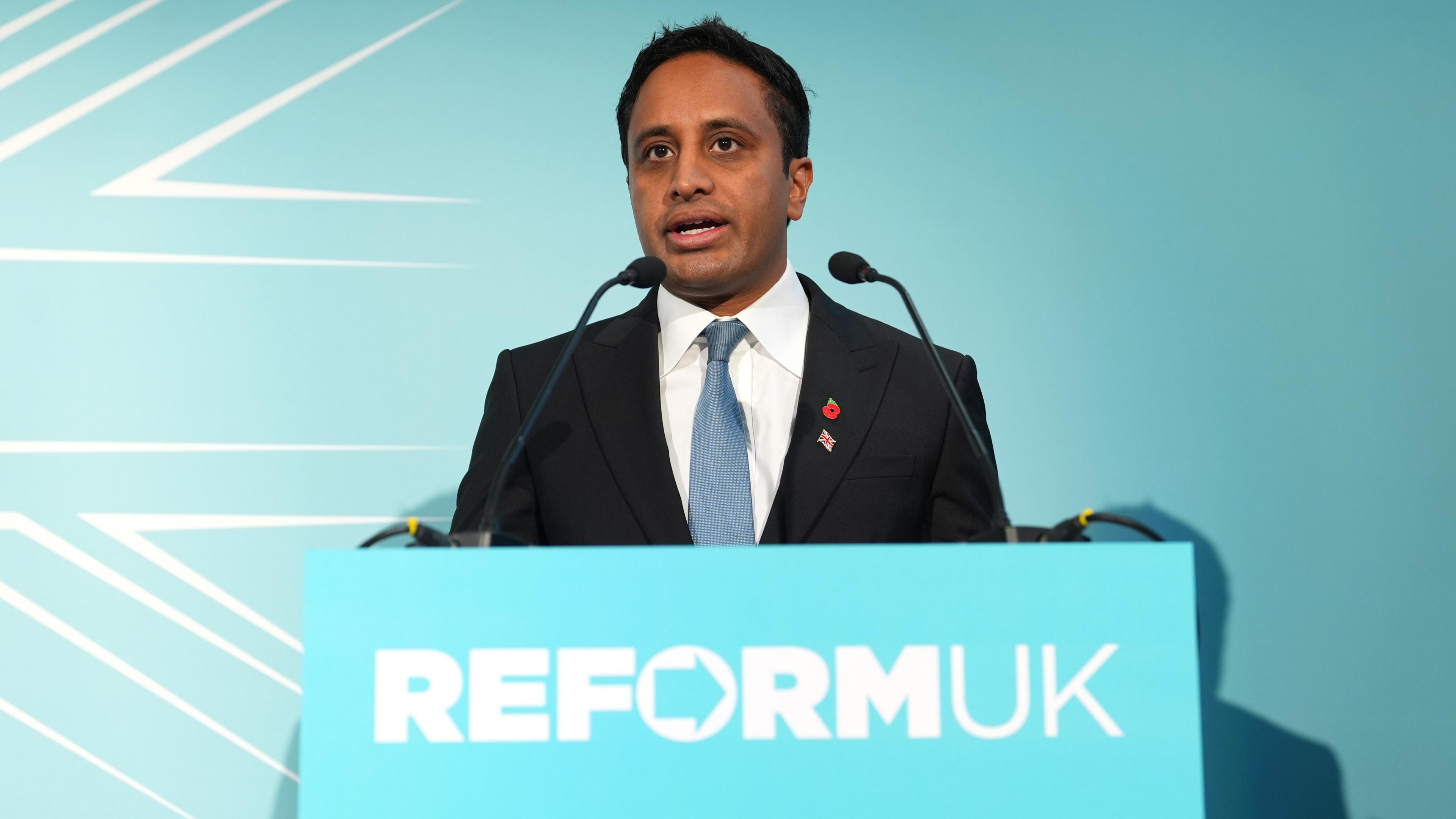 Head of Policy Zia Yusuf speaking during a Reform UK press conference at Church House Westminster in London. He has short black hair and is wearing a dark navy suit with a white shirt and light blue tie. He wears two small pins on his lapels - a poppy and a Union Jack flag. He stands behind a podium bearing a light blue sign that reads Reform UK. 