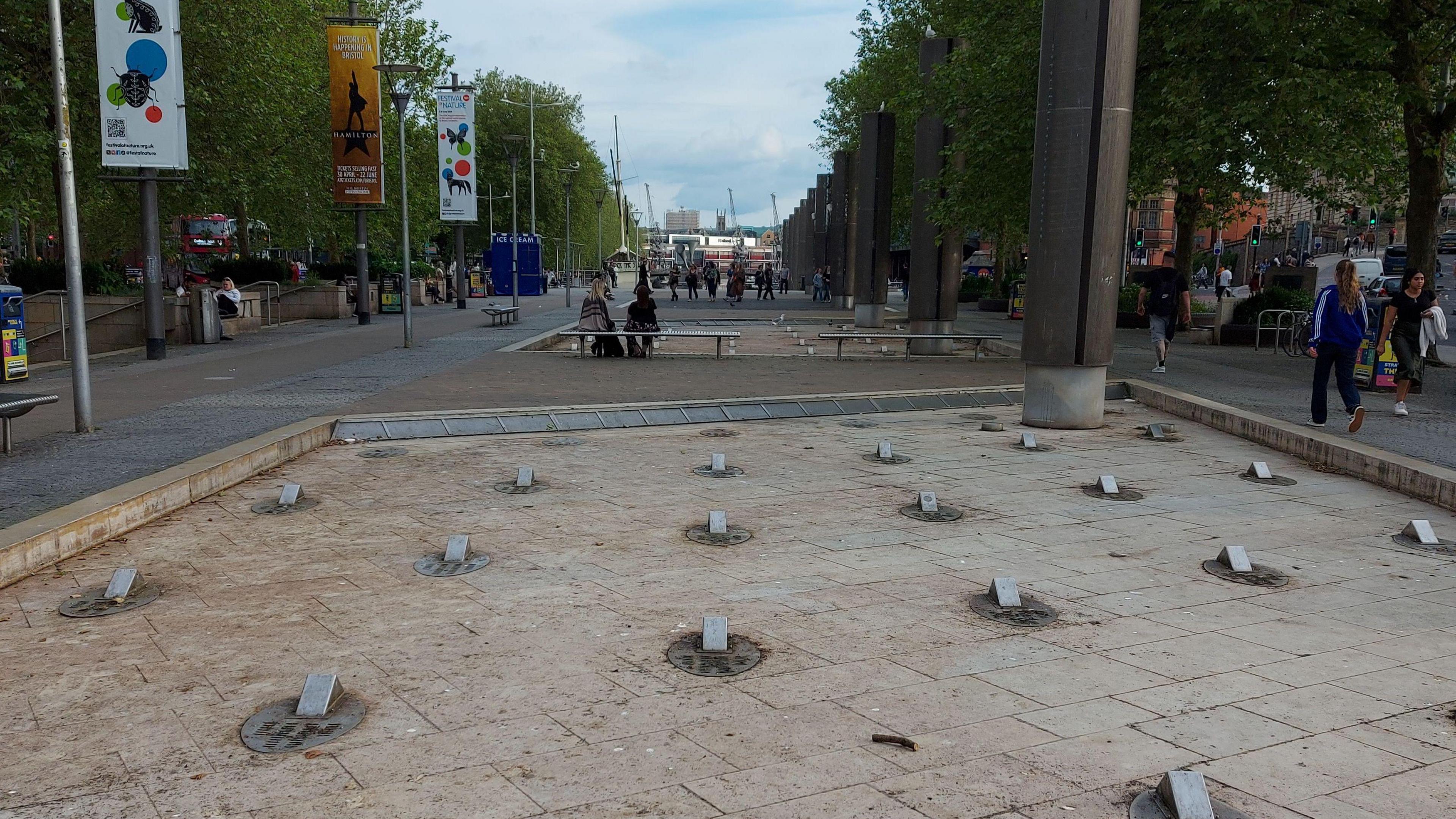 Bristol city centre promenade with fountains that are off in the foreground. These people walking around and signs in the walkways.