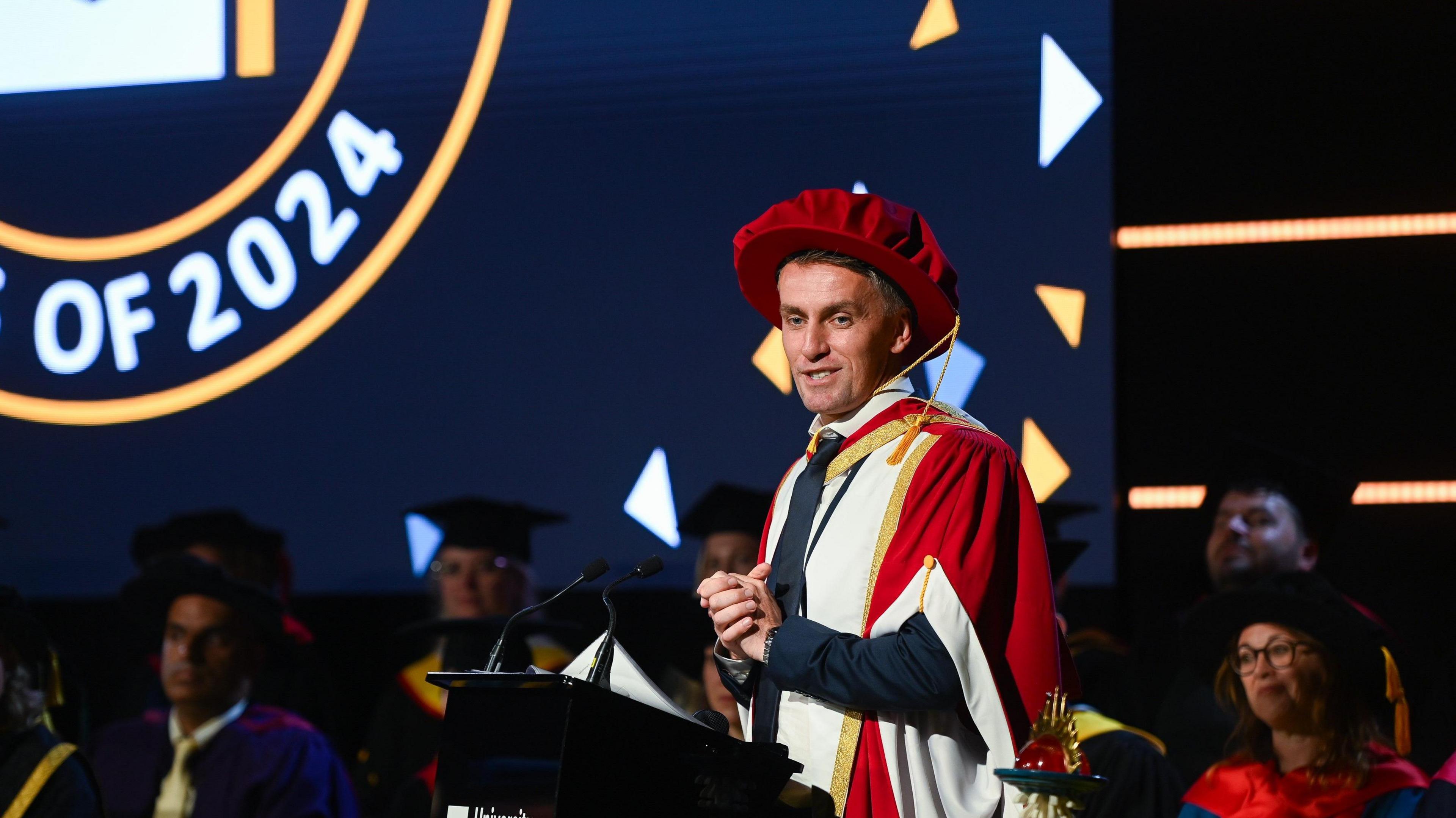 Kieran McKenna wearing a dark suit and red academic gown and hat standing at a lecturn with hands clasped. Behind him are blurred images of people wearing academic robes.