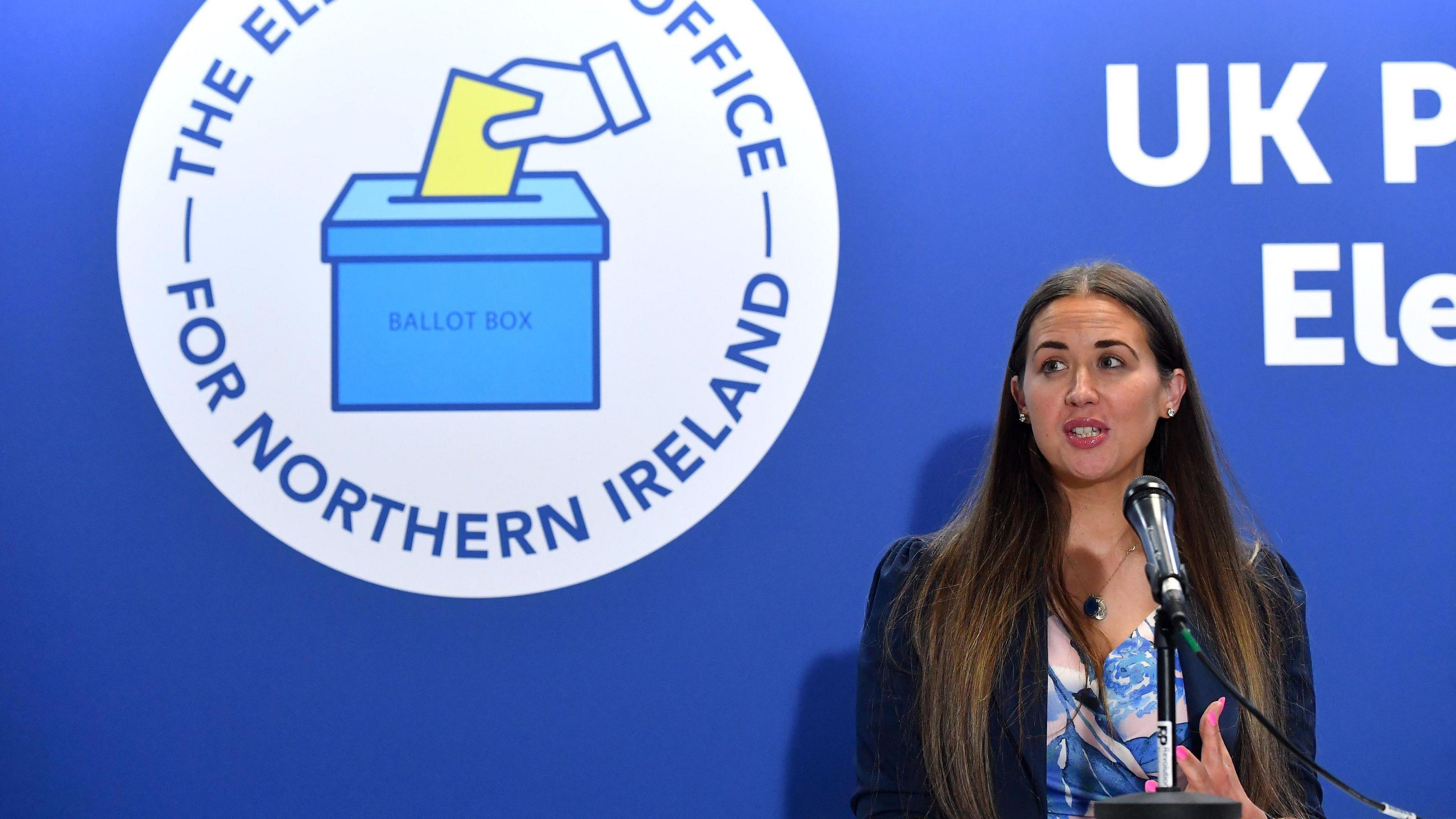 Sorcha Eastwood, right, standing in front of a blue wall with the Electoral Office logo on it to the left 