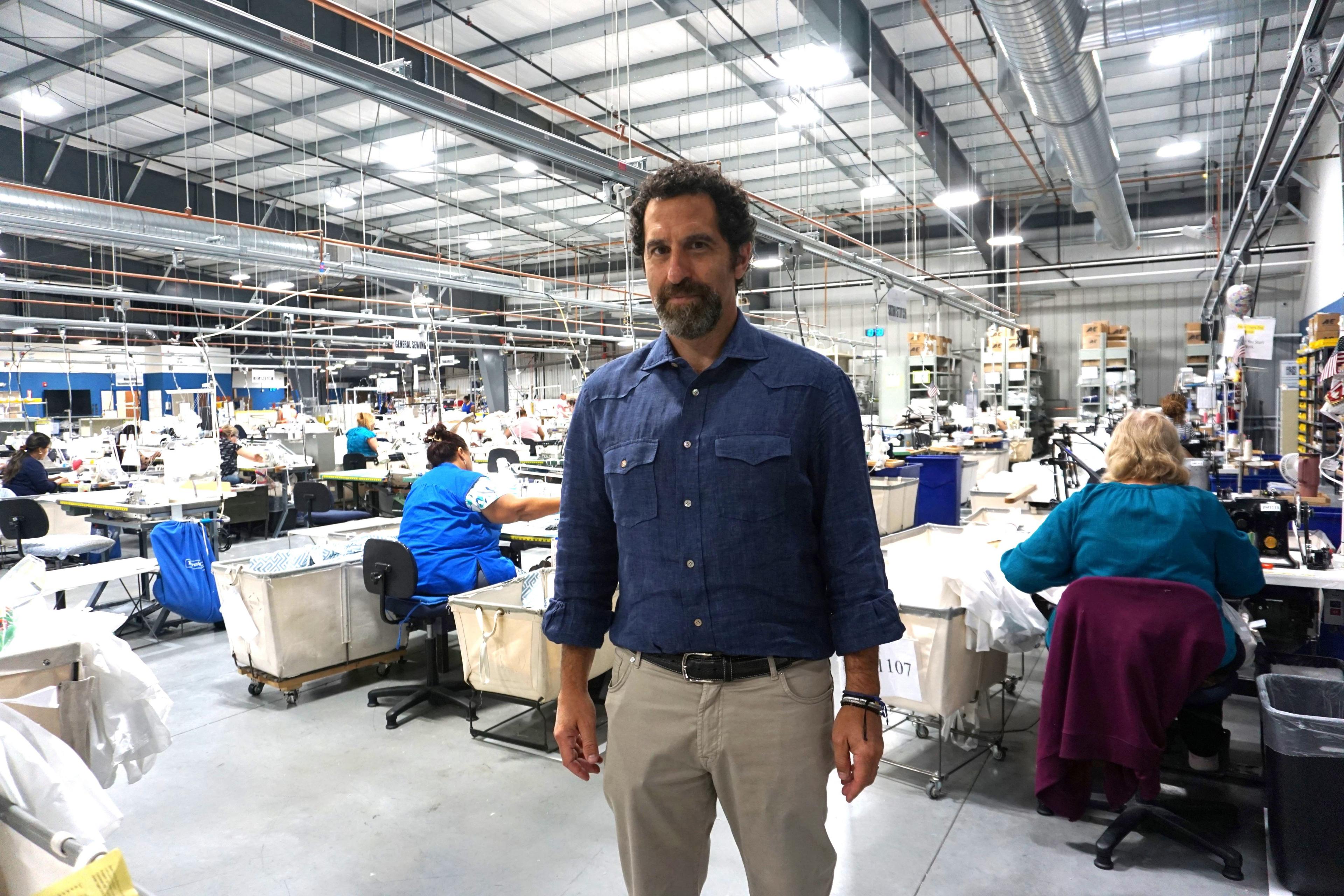 George Matouk, in a blue button down shirt, at his factory in Fall River. Behind him women are seated at workstations in the large warehouse space.