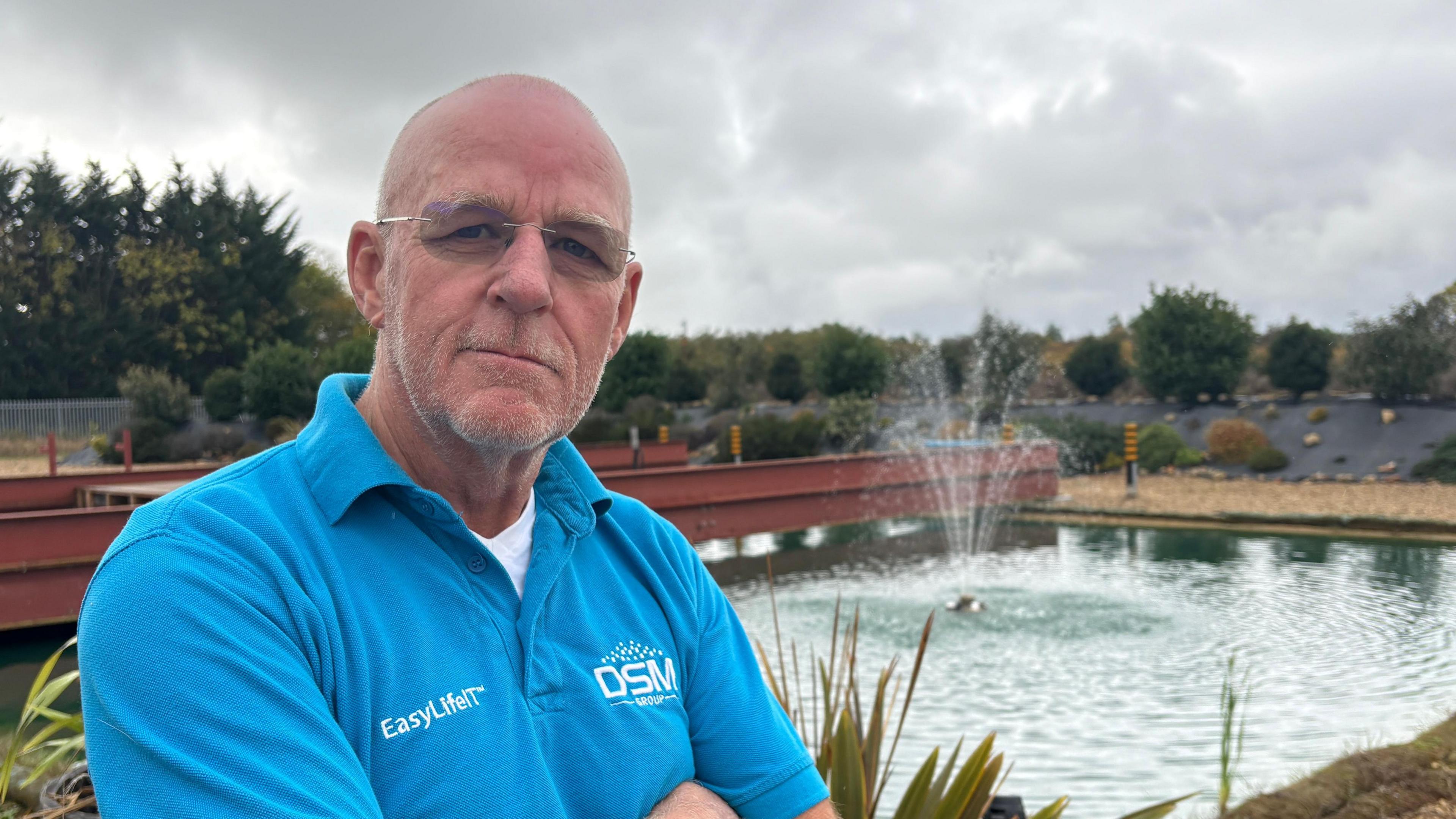 Mike Richardson looking directly down the camera and standing in front of a rectangular lake outside. His arms are folded and he is wearing a blue collared t-shirt, with EasyLifeIT and DSM logos on either side of his chest. He appears mostly bald but has short, grey stubble on his face. He is wearing partially tinted glasses. Behind him is a lake, with a small fountain spraying jets of water about two metres into the air. In the further distance are trees. The sky looks cloudy and grey.