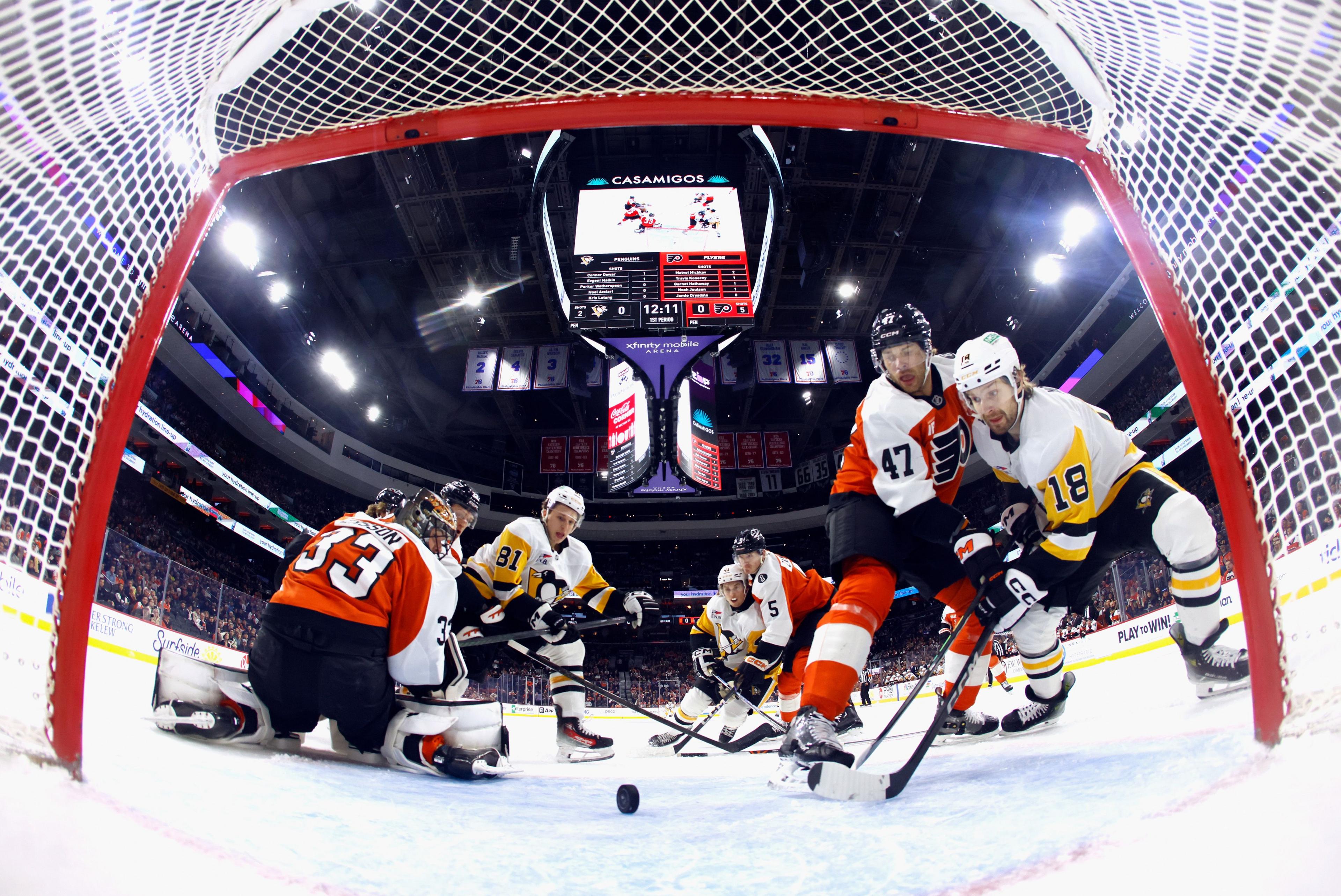 View from inside a hockey goal showing players from two teams battling for the puck near the crease. The goalie in orange and black kneels while others crowd the net. Overhead scoreboard and bright arena lights dominate the background.