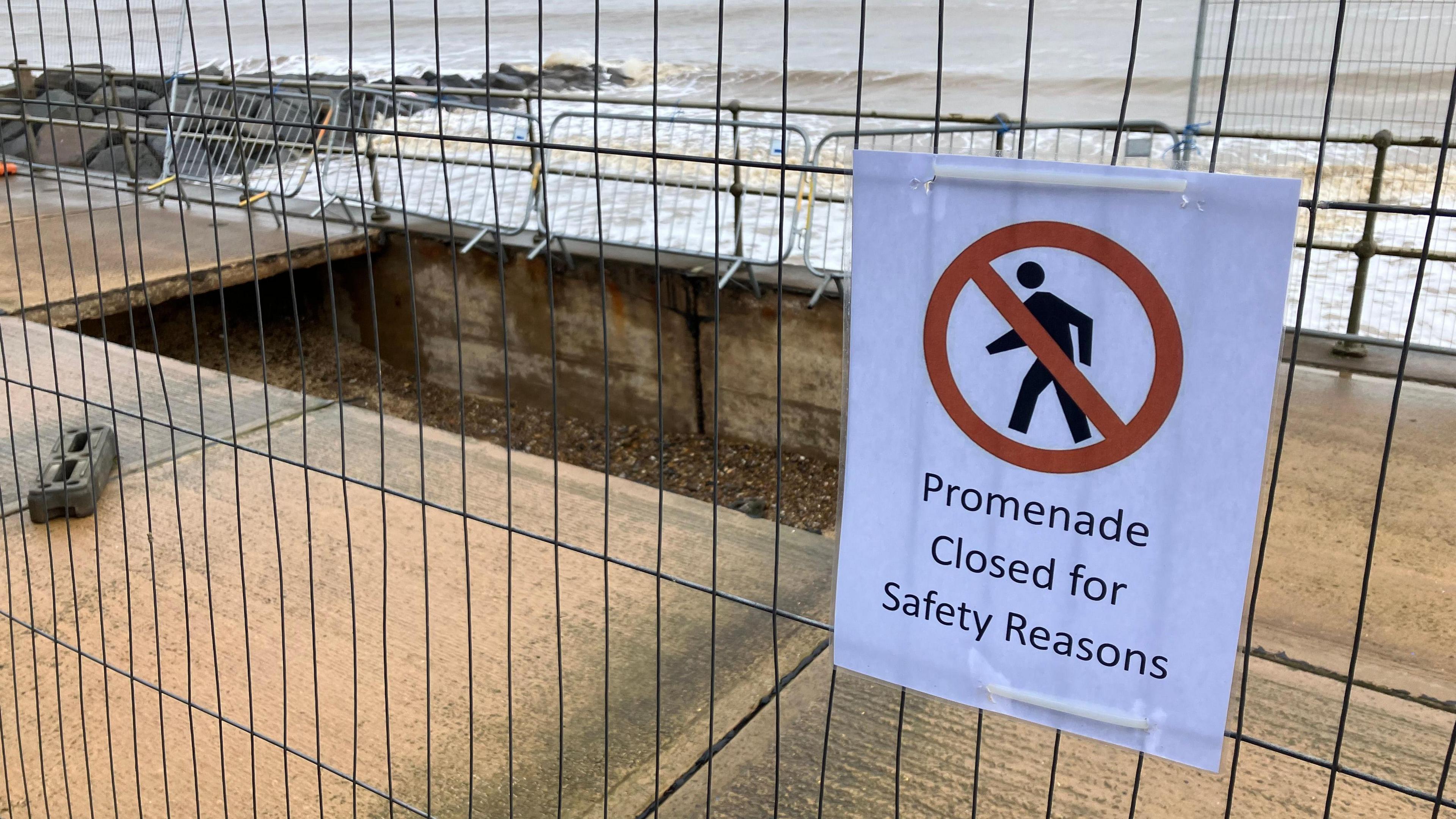 Metal fencing surrounds a large, rectangular-shaped hole in a concrete promenade with a sign on the fence reading "promenade closed for safety reasons". Waves and a rock groyne can also be seen in the background. 