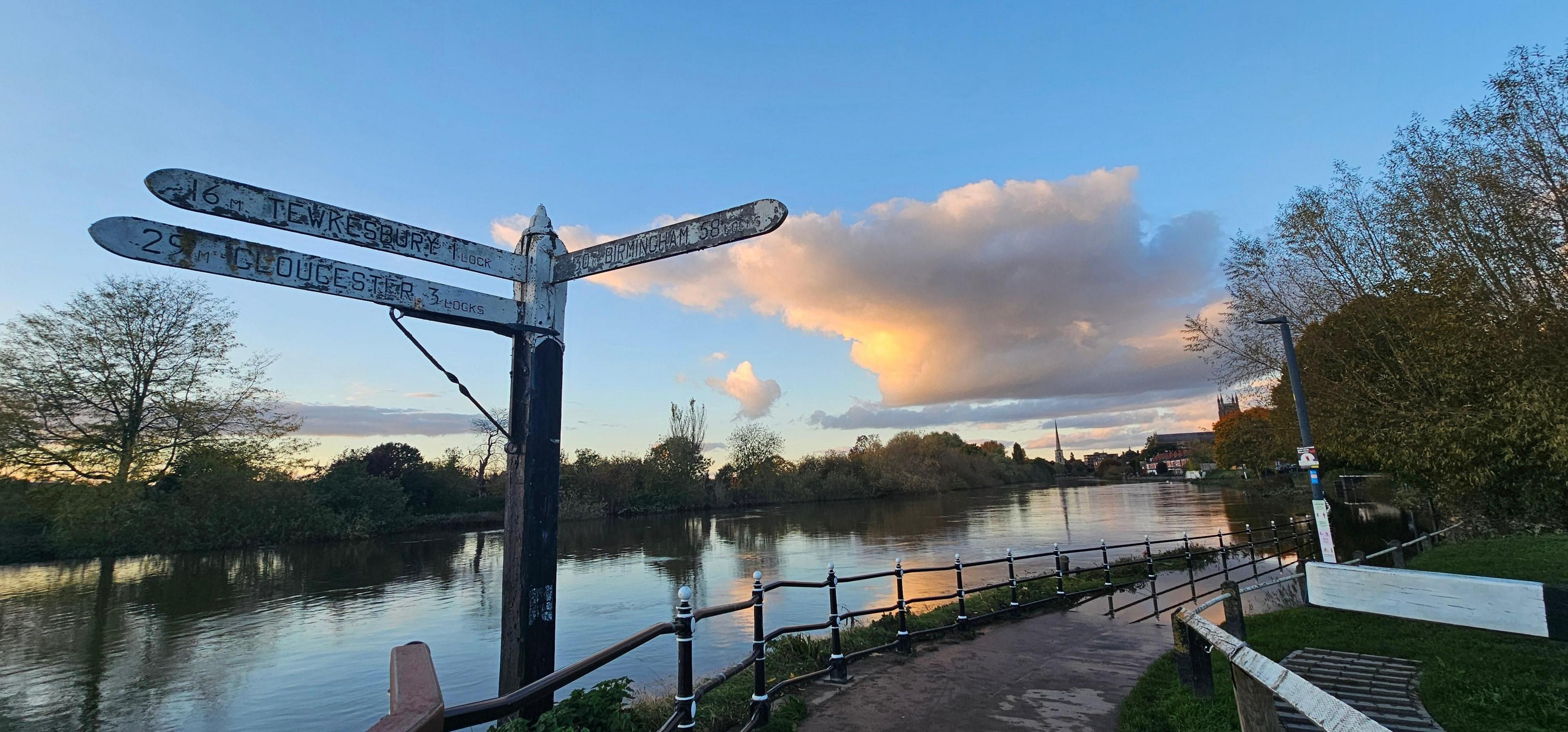 A faded wooden sign showing Tewkesbury and Gloucester in one direction, heading south on the river, and Birmingham north east along the canal. The river is in the background, reflecting a blue sky with white clouds.