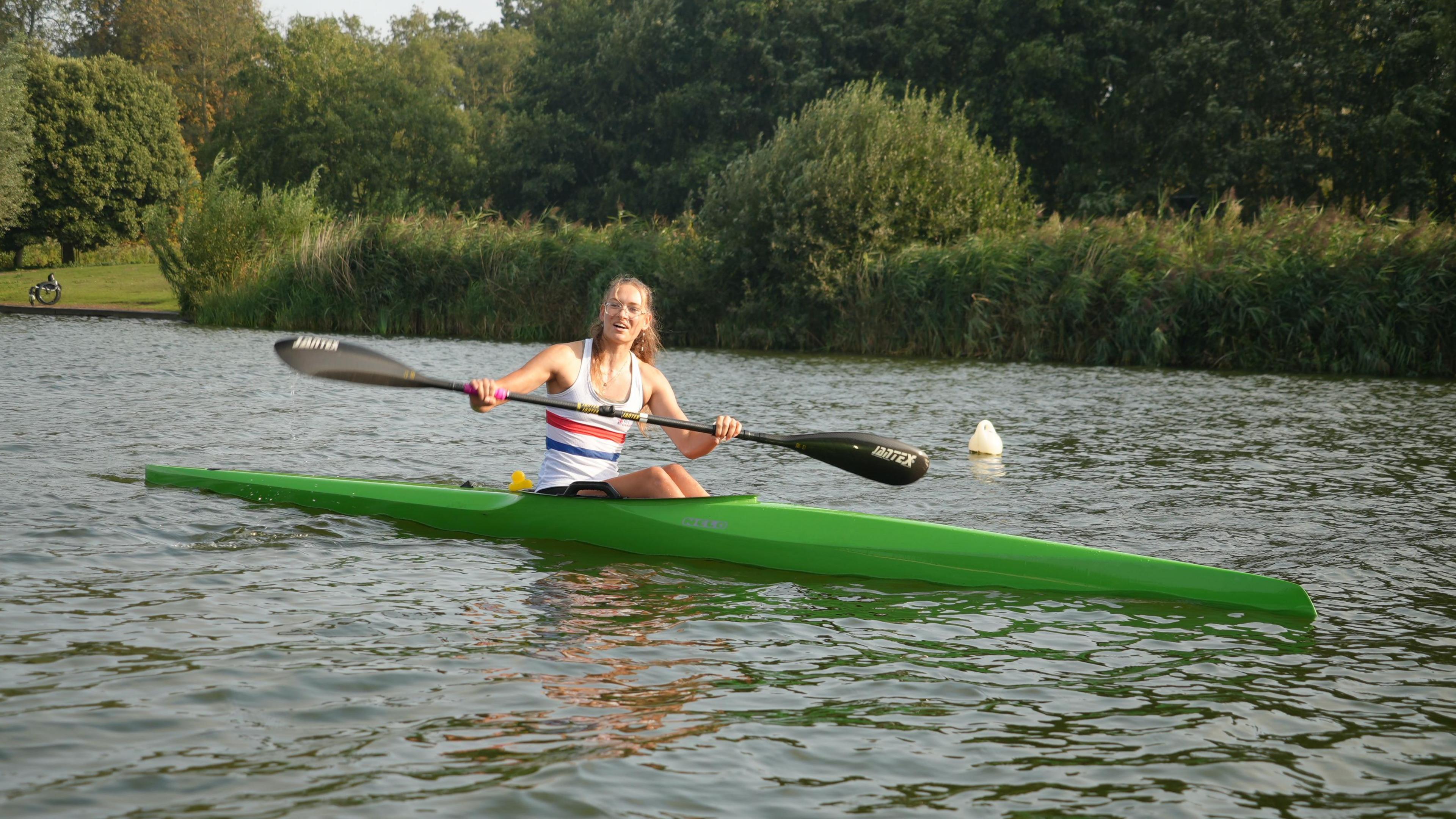 Kamila Sklenarova in a green canoe boat smiles at the camera with her paddle raised, on a body of water with greenery in the background. 