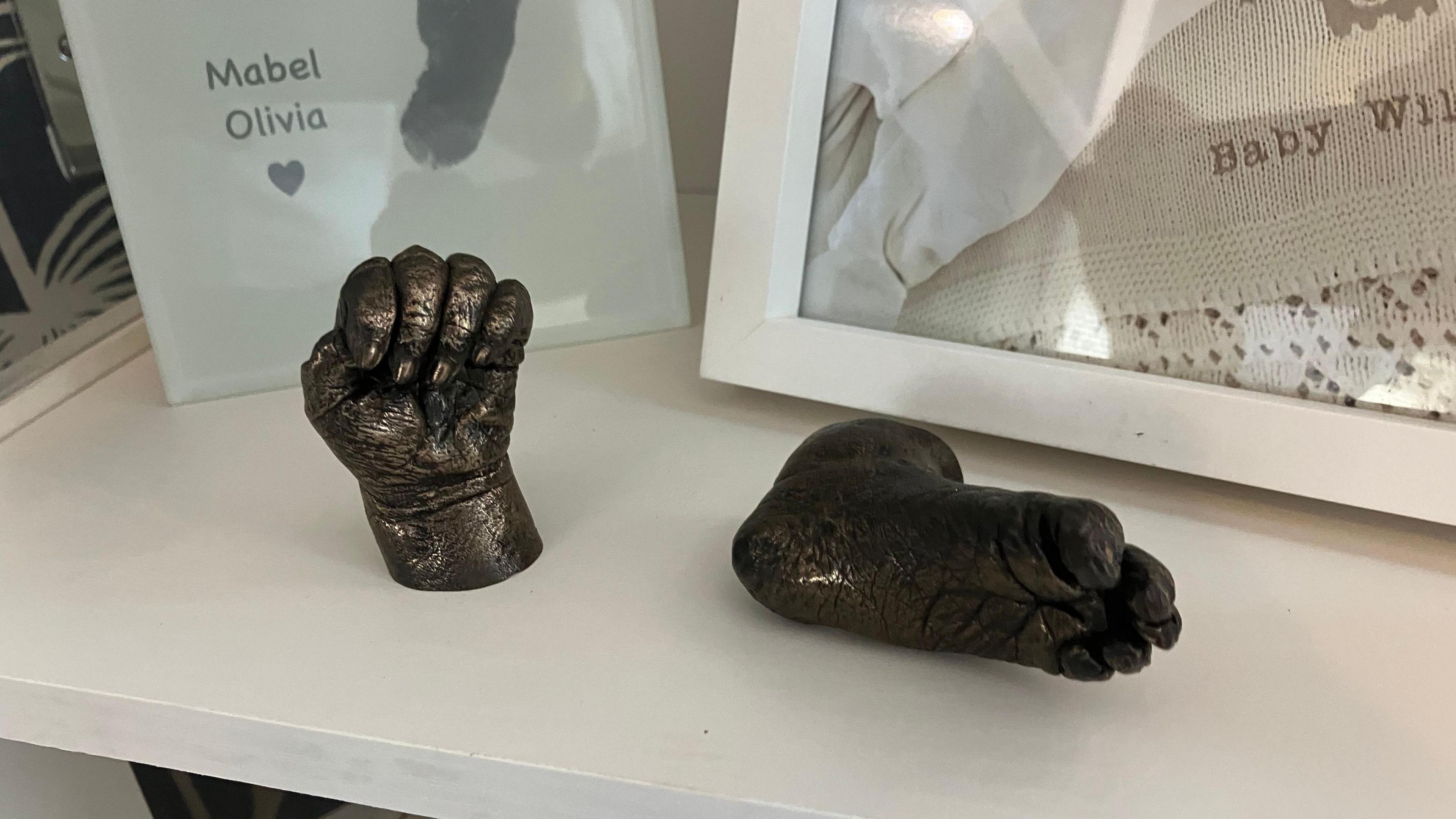 A small set of dark grey castings were made of Mabel's hands and feet. They are placed in a small white cabinet, in front of various other items to remember Mabel such as a printed footprint of her and a framed picture of her.