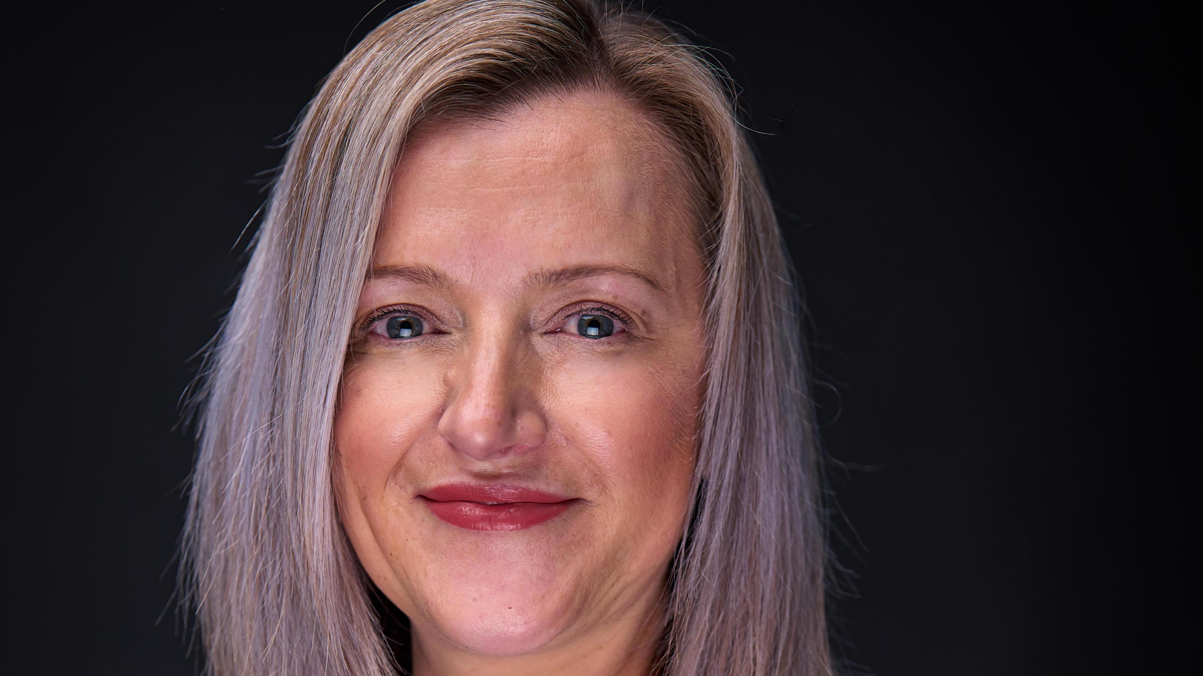 A blonde woman's face smiling at the camera. She has blue eyes and is wearing red lipstick