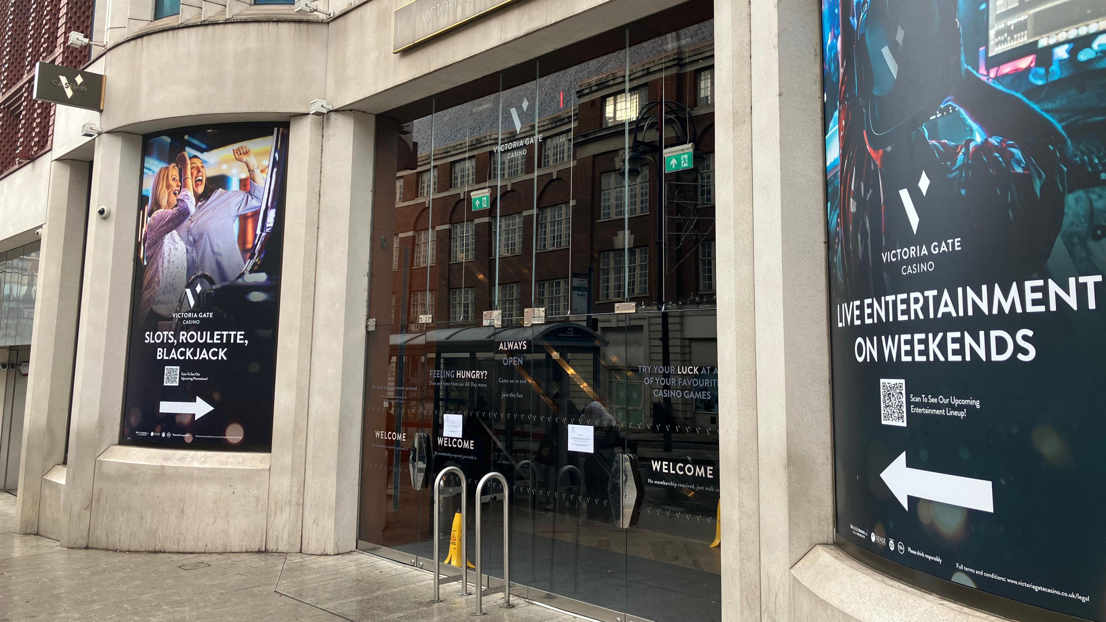 Street view of the entrance to Victoria Gate Casino. The building has large glass doors with reflective surfaces showing nearby architecture. On the left side, a poster advertises ‘Slots, Roulette, Blackjack’ with an image of two people enjoying drinks. On the right side, another poster promotes ‘Live Entertainment on Weekends’ featuring a futuristic figure in neon colours.