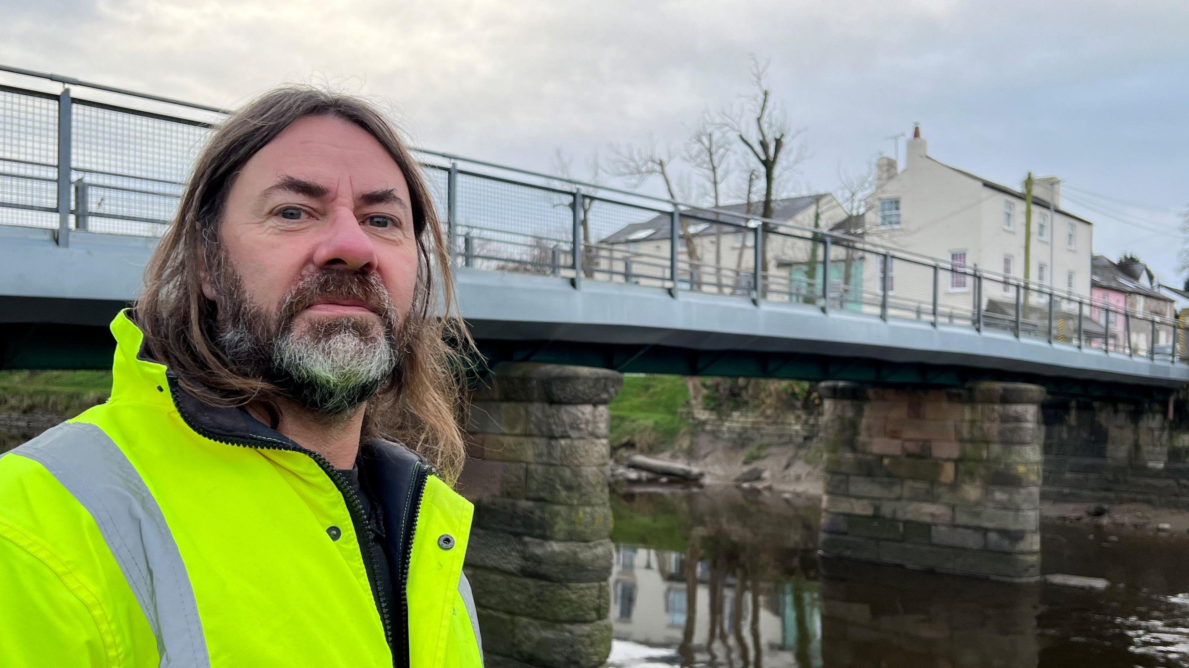 Steve Houghton who has long brown hair, beard and moustache wearing hi-vis jacket standing in front of the toll bridge which crosses the River Wyre