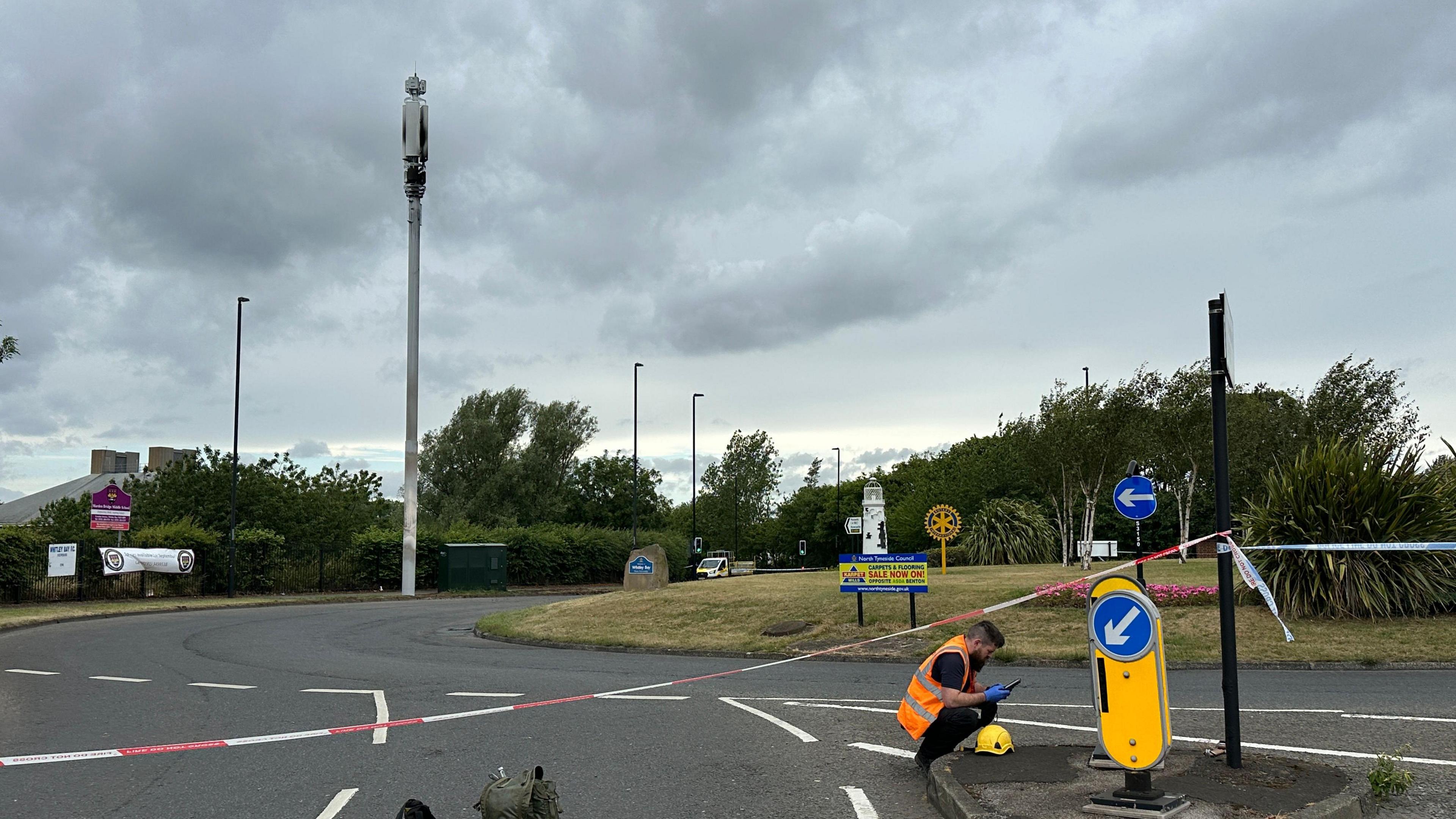 Burned white telephone mast next to roundabout with grass on it. Surrounding it are police tape and a man with a high-vis orange jacket checking something on the ground