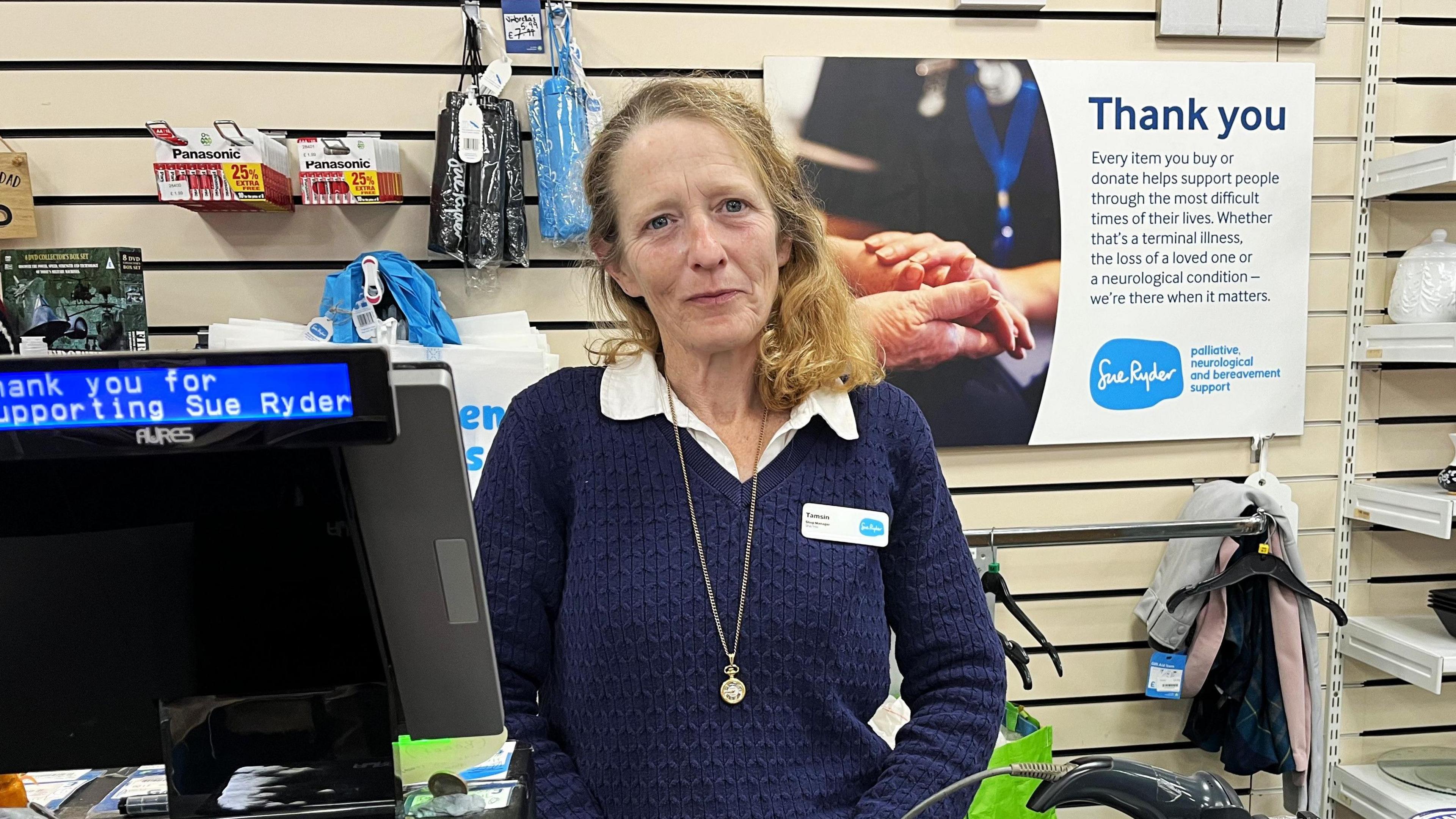 Tamsin Payne is wearing a dark blue jumper on top of a white, collared shirt. She's also wearing a necklace with a pendant and a name badge. She's standing behind the till of her shop.