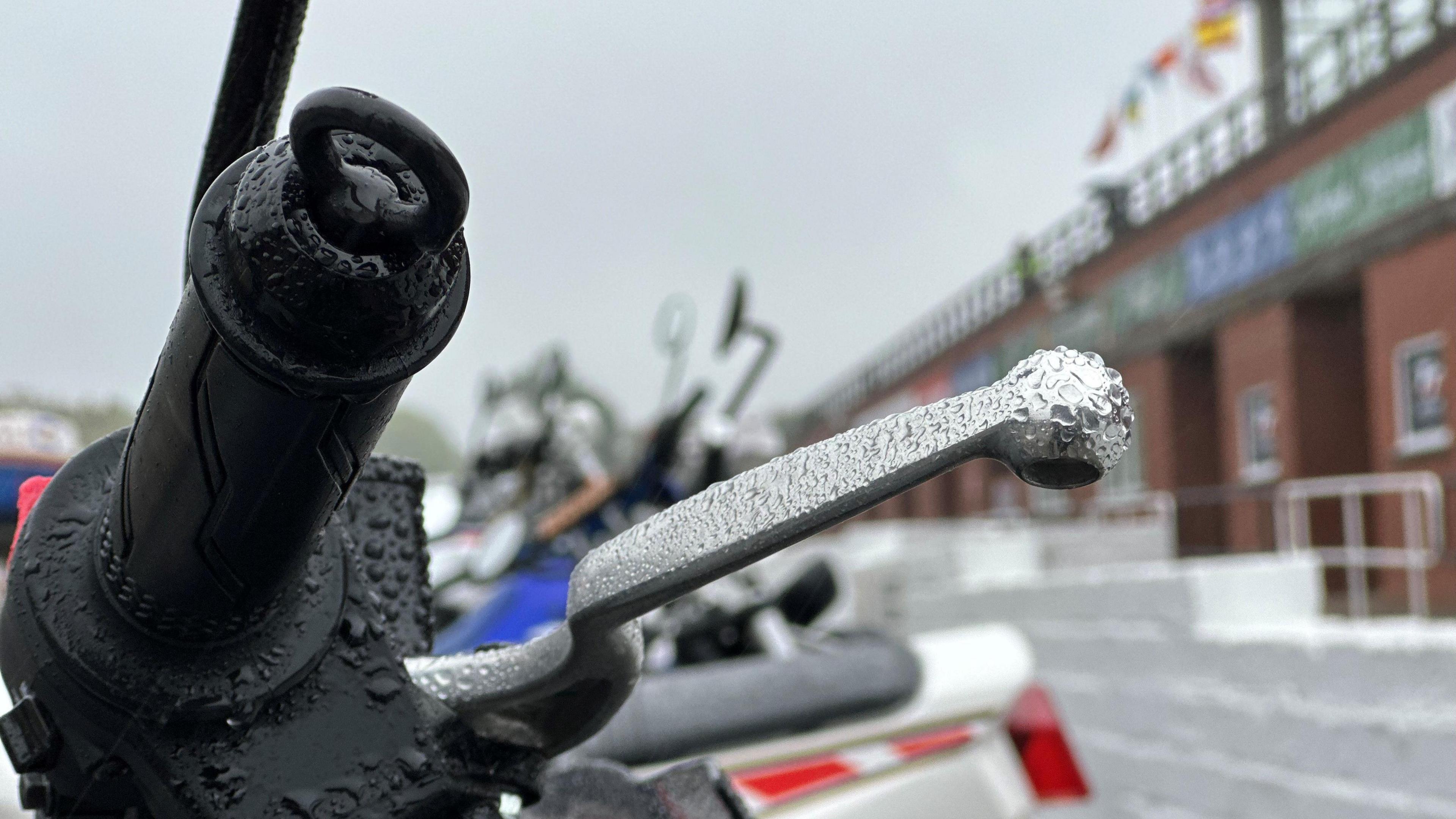 The black handle and silver brake lever of a motorcycle with droplets of rain gathered on them. A line of motorcycles parked in front of the grandstand can be see in the background.