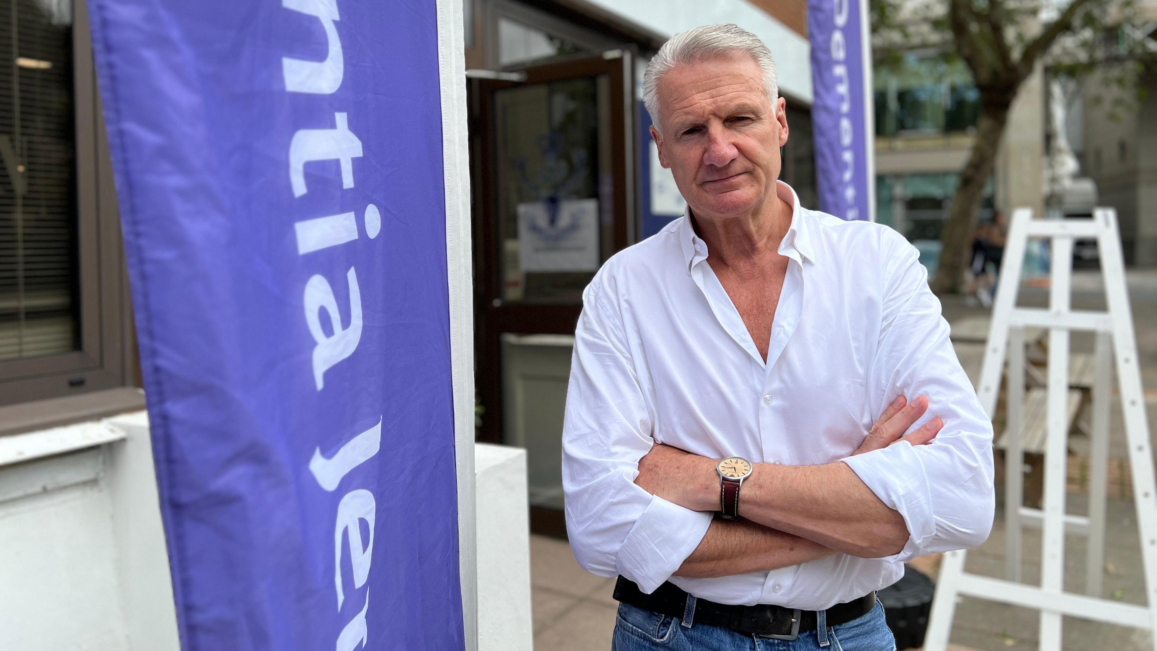 Deput Tom Binet looks at the camera with his arms crossed outside by a Dementia Jersey flag