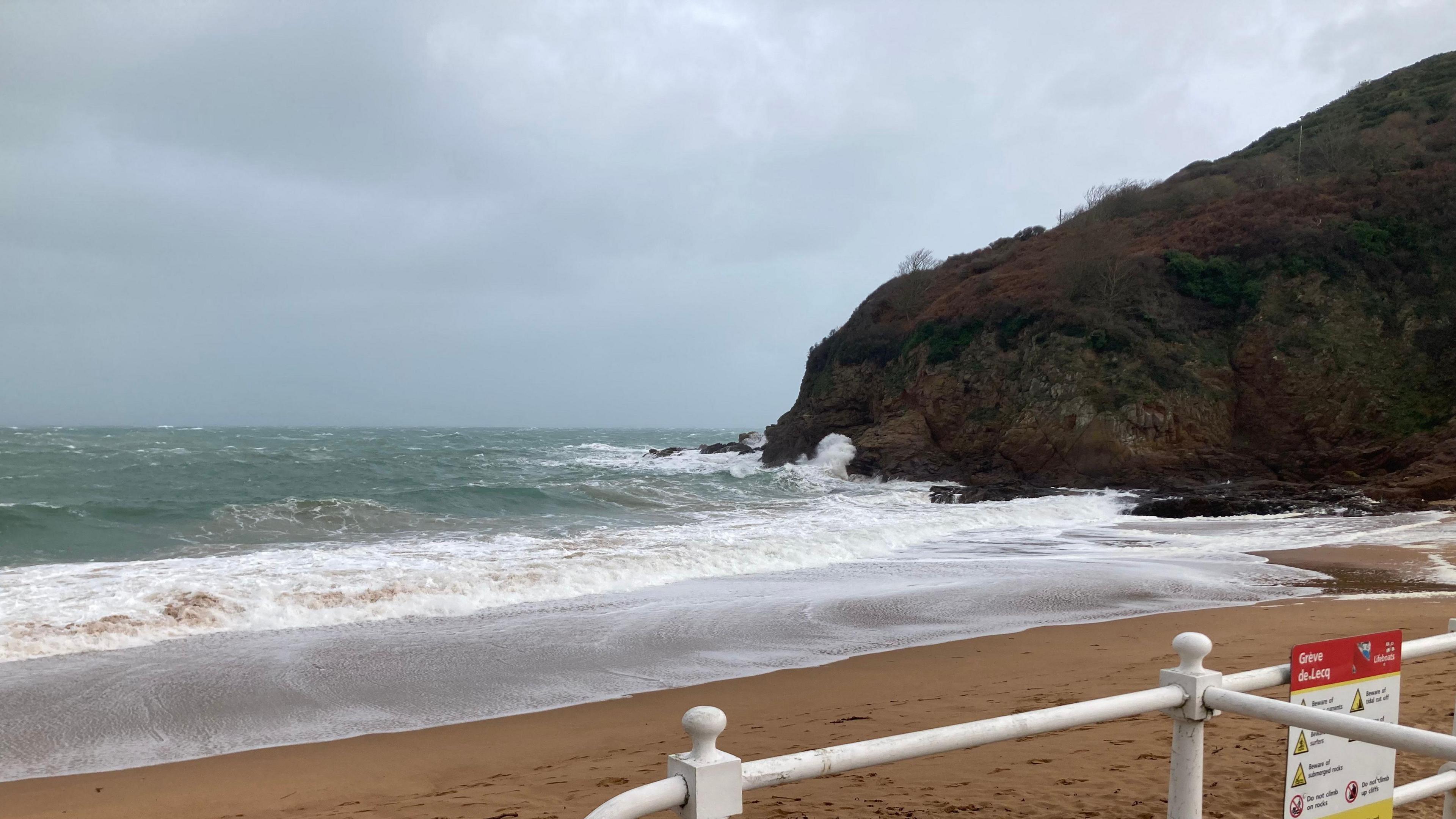 Jersey Grève de Lecq beach. Waves are crashing against the brown sand. There is a cliff on the right and white railings in the forefront.