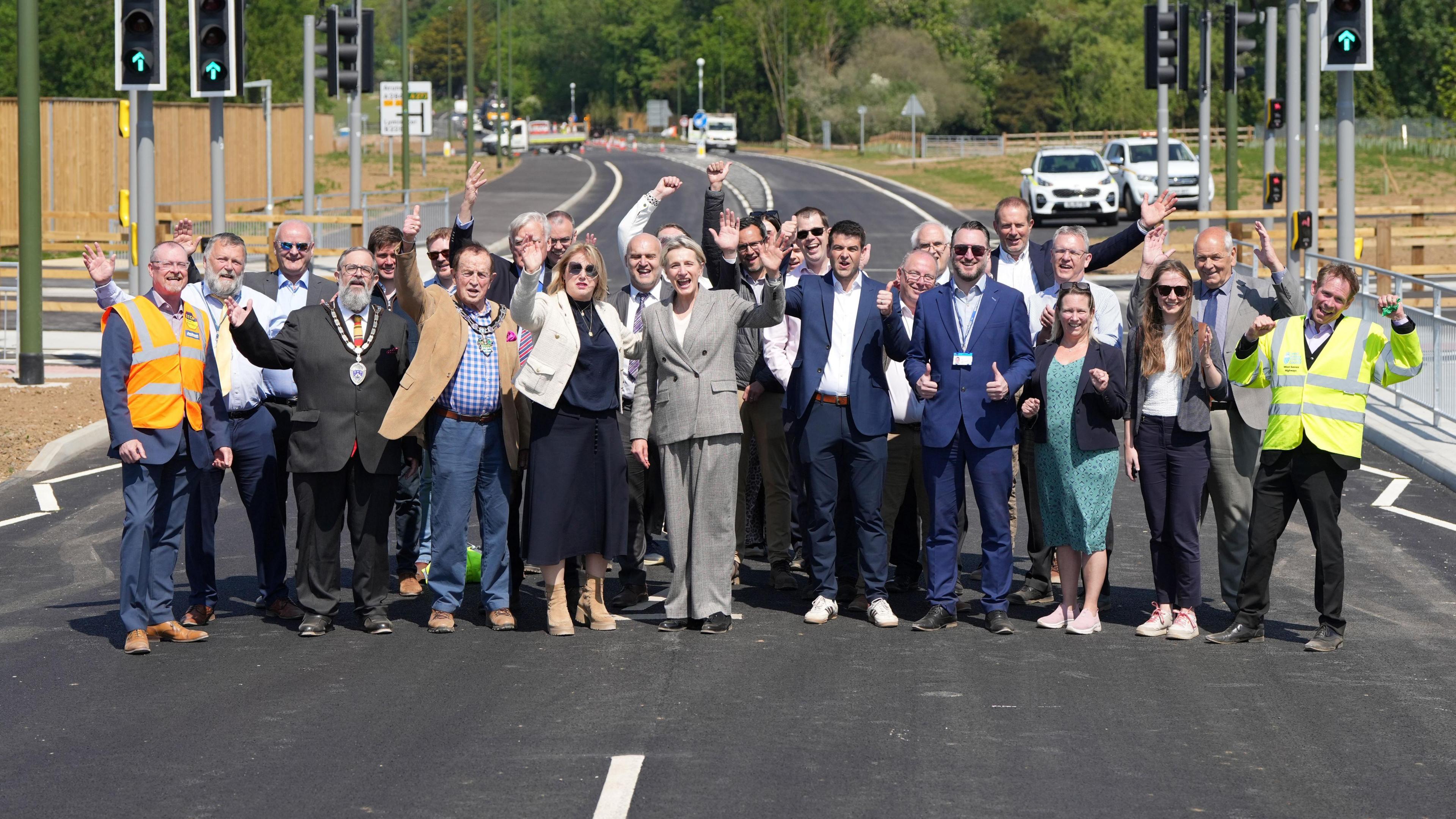 A group of people stood in the middle of a road. They are holding their hands up and holding their thumbs up to the camera while smiling.
