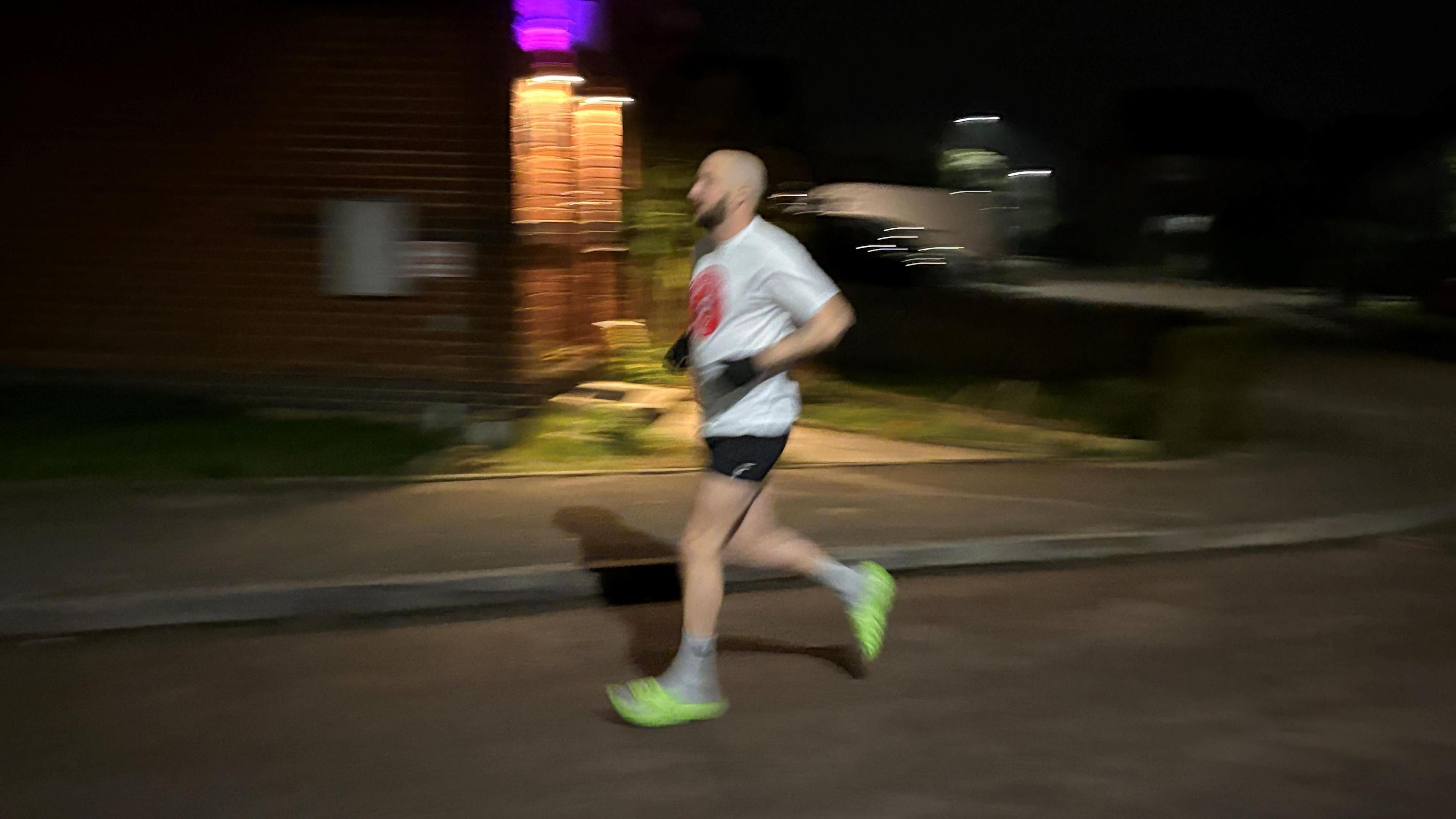 Daniel Fairbrother running in a street, in the dark, he is wearing bright coloured sliders, black shorts, white T-shirt and black gloves. He is blurred due to running. 