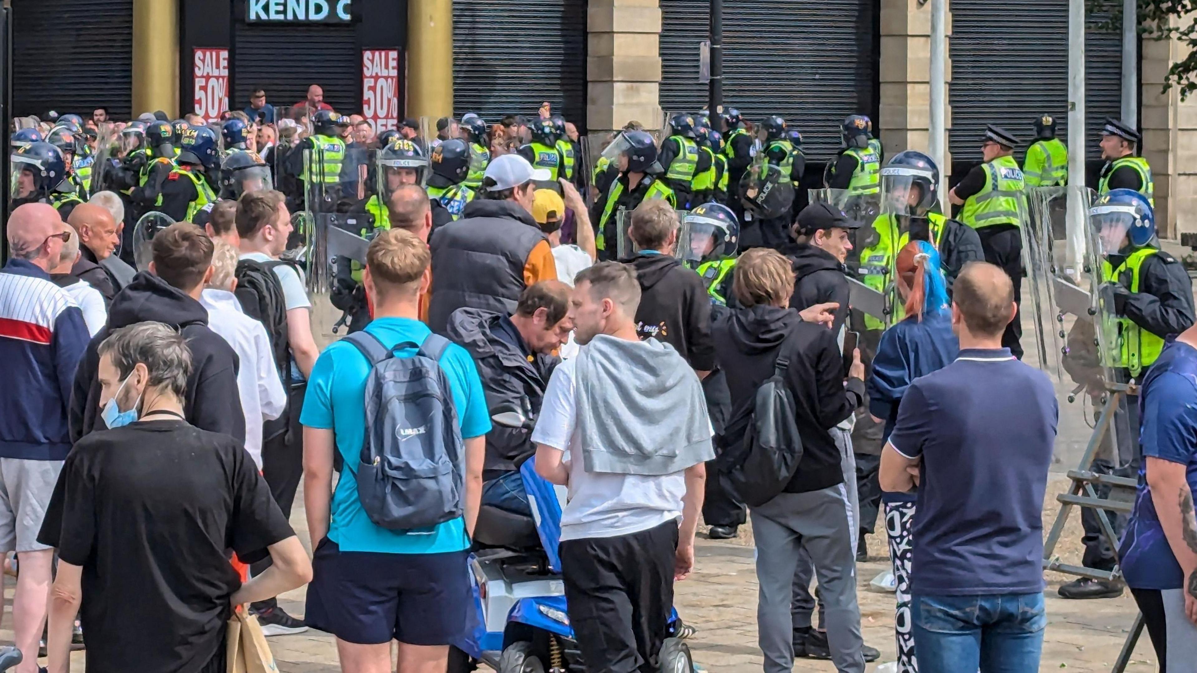 Crowds of people standing in front of police officers holding riot protection gear