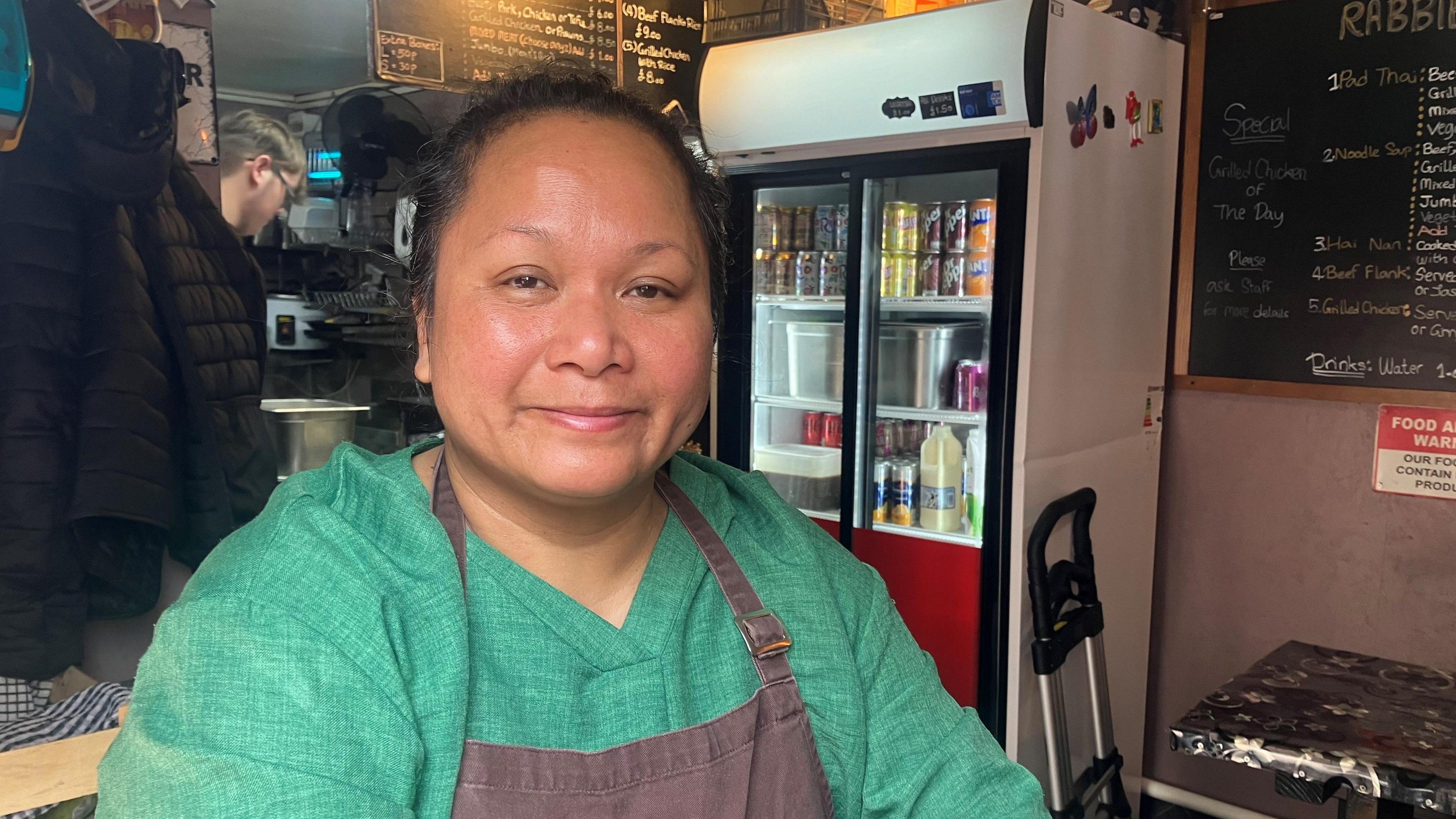 Rabbe Promat in her Thai takeaway. She's wearing a green, linen, long-sleeved top and a brown apron. Her dark hair is tied back and she is standing in front of a menu board and a drinks fridge