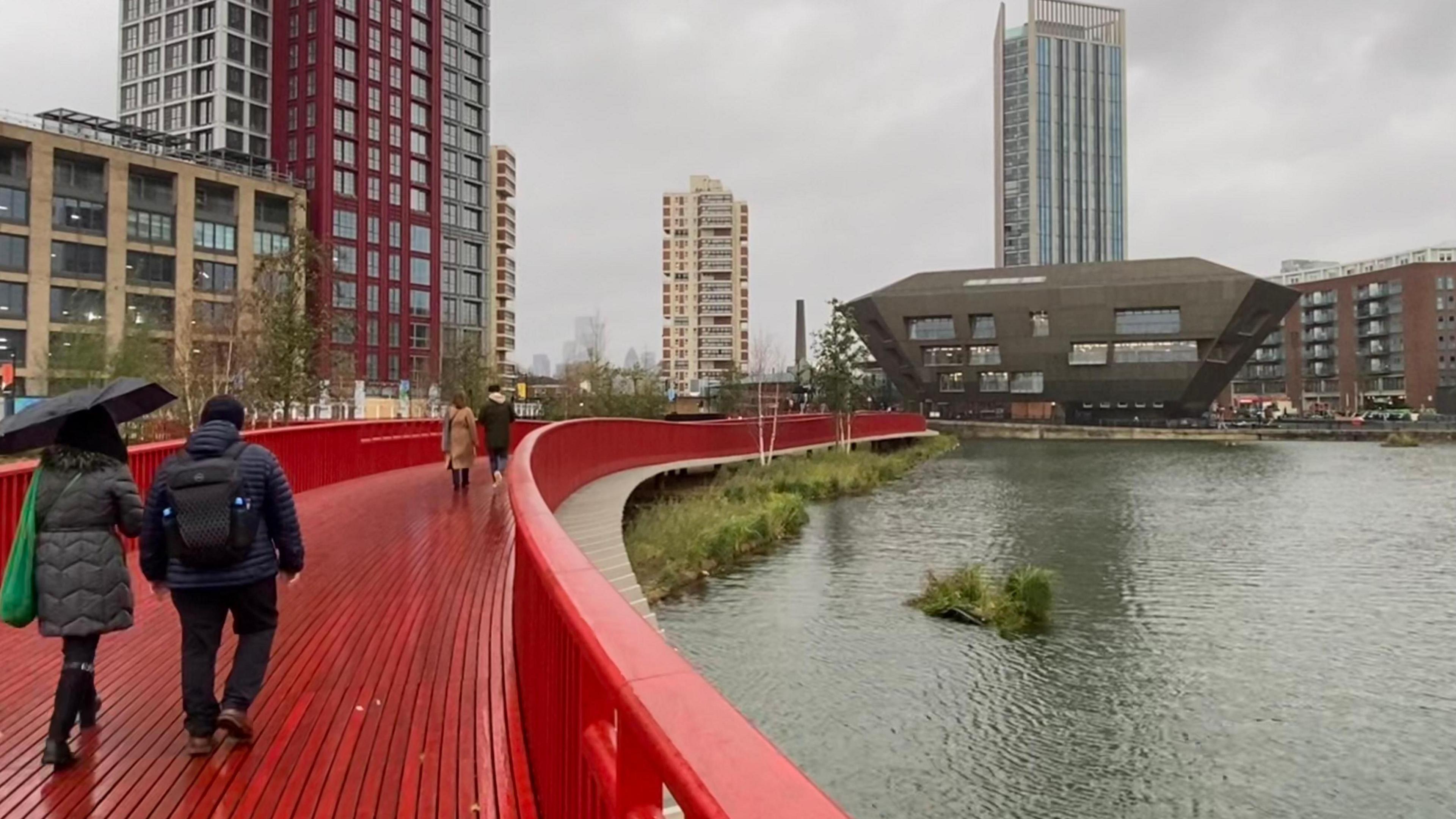 Grey skies in London, two people are seen sheltering under an umbrella as they walk over a red bridge